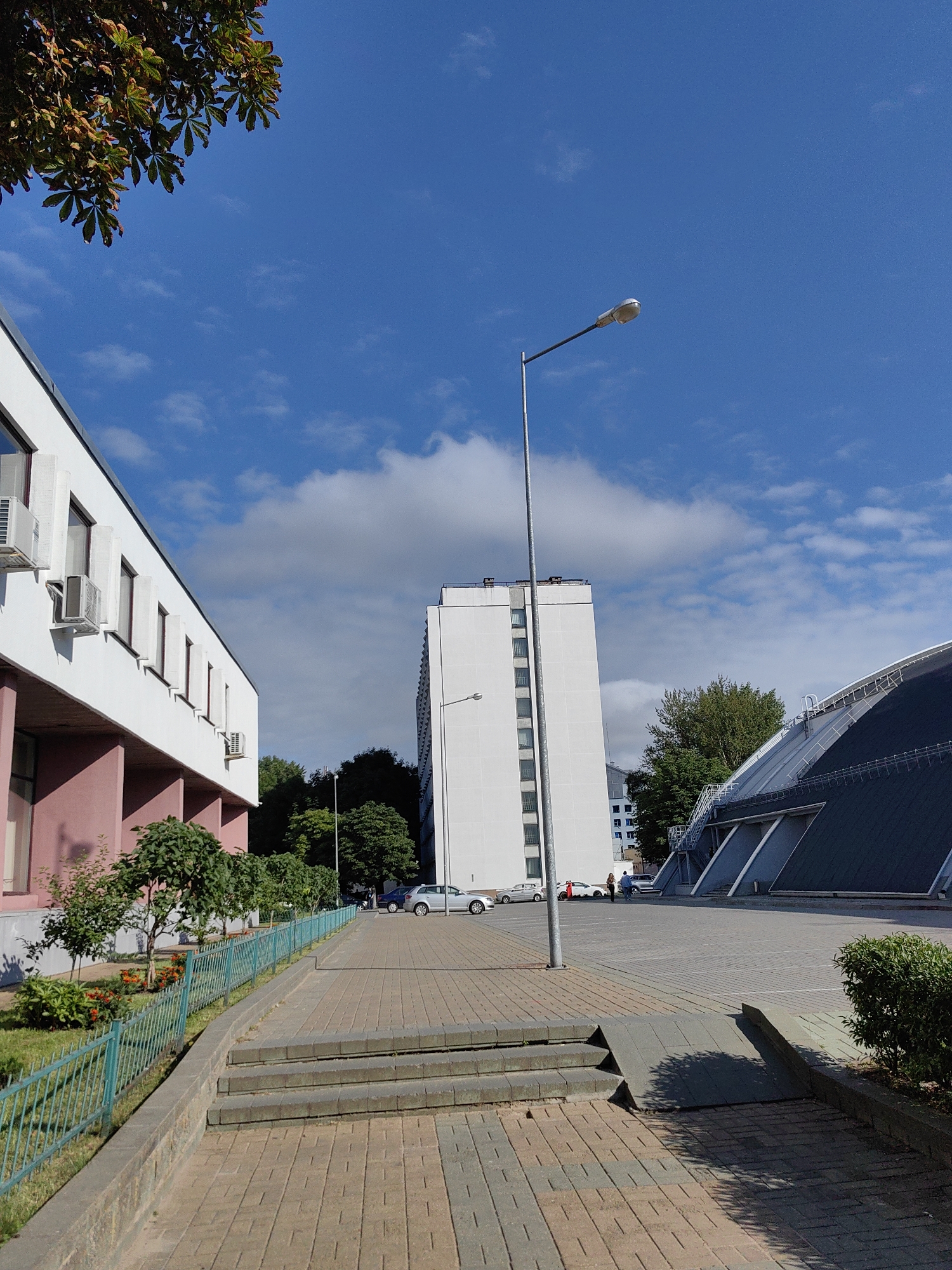 The image shows an outdoor scene on a sunny day with a bright blue sky and scattered white clouds. In the foreground, a paved walkway with brick-like patterns leads towards a set of concrete steps and a ramp. A low, turquoise metal fence borders a grassy area with small trees and orange flowers on the left side. Behind the walkway, a white building with pink accents is visible, featuring windows with visible air conditioning units. Further in the background, a tall, white, rectangular building stands against the sky. To the right, a modern building with a dark, sloping roof structure is partially visible. Several cars are parked in a lot between the buildings. A single street lamp with a white fixture on a tall pole stands near the center of the image. In the far distance, more trees and buildings are visible. A branch of a tree with green and brown leaves hangs into the top left corner of the frame.