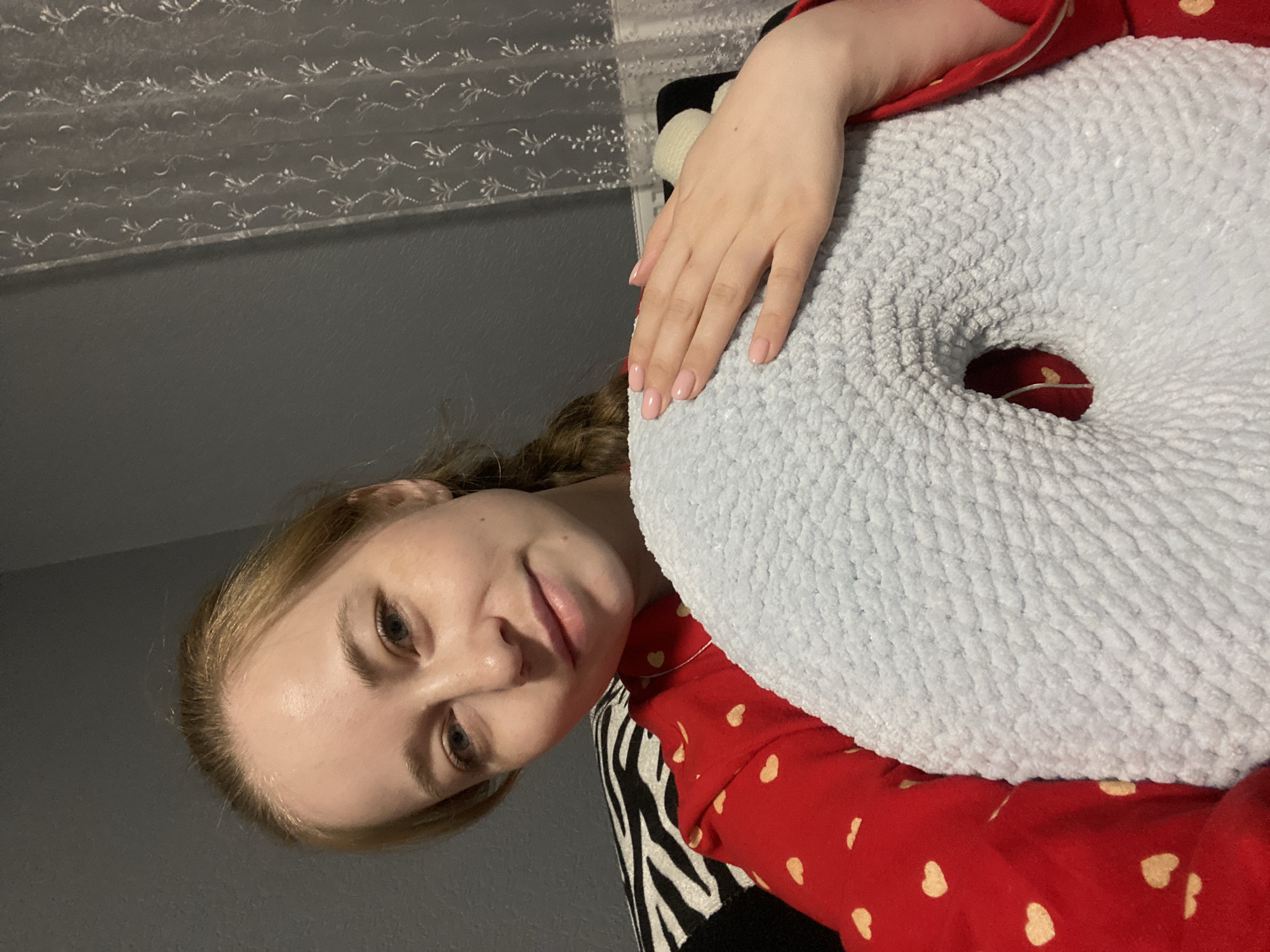 A young woman is captured in a close-up, indoor portrait, likely taken in Saint Petersburg, Russia. She is lying down, facing the camera with a gentle, slightly smiling expression. Her light brown or blonde hair is pulled back, revealing her forehead.
She is wearing a red top or pajamas patterned with small, light-colored dots or hearts. Her left arm rests upon a large, light blue, plush, donut-shaped pillow that occupies the right side of the frame. Her hand, adorned with light pink nail polish, is visible on the textured surface of the pillow. The central hole of the pillow reveals more of her red attire. Partially visible beneath her arm is a black and white zebra-print fabric, possibly part of another pillow or bedding.
In the background, a plain dark gray wall meets a lighter-colored curtain or window treatment featuring a delicate, repetitive white floral or vine pattern. The lighting is soft and appears artificial, suggesting an evening or nighttime setting. The overall atmosphere is relaxed and personal. No text is visible in the image.