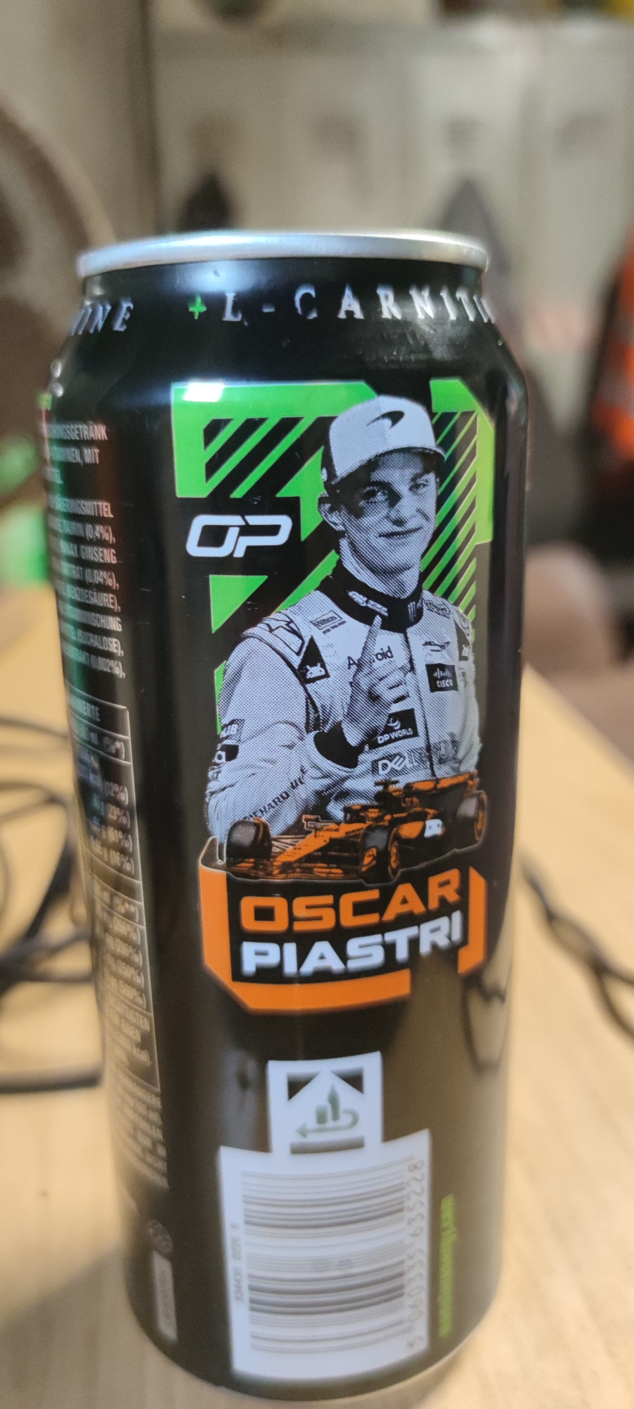 This is a close-up, slightly angled shot of a black energy drink can. The can features a prominent image of a young man in a racing suit and cap, identified by the text "OSCAR PIASTRI" below him. He is giving a thumbs-up, and a Formula 1 car is depicted in front of him. The can also has green accents and text, including "OP" and what appears to be "L-CARNIT." Nutritional information is visible on the left side of the can, with some text in German, as indicated by "INKUNGSGETRANK" (Energy Drink). A barcode is visible at the bottom right. The background is blurred, but suggests an indoor setting, possibly a desk with some cables. The lighting appears to be artificial, casting some reflections on the can. The overall impression is that of a promotional product related to a motorsport athlete.