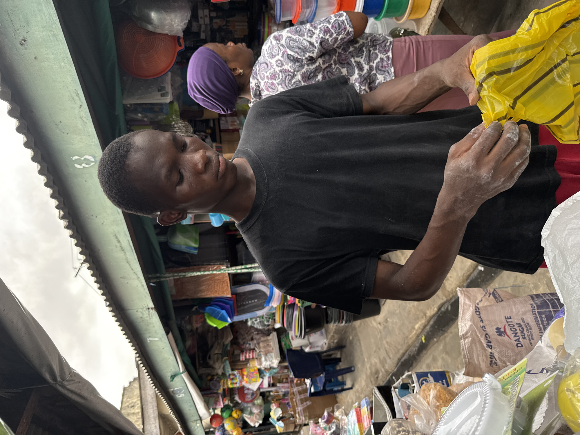 This image, captured in Lekki, Nigeria, depicts an active market stall during daylight hours. The scene is slightly rotated clockwise from an upright view.
In the foreground, a young person with dark skin and short dark hair is engrossed in an activity. They wear a black short-sleeved t-shirt and red shorts, partially visible at the bottom right. Their hands, notably covered in a white powdery substance (possibly flour or sugar), are holding and manipulating a yellow plastic bag with dark stripes. This suggests they are in the process of packing, weighing, or portioning goods. Adjacent to their hands, several packaged items are visible, including clear plastic bags containing what appear to be food products like bread or pastries, and various white plastic dishes. A white paper bag clearly displays the text "DANGOTE SUGAR" and "INFORMATION," providing a strong local context.
Behind the young person, an adult woman is visible. She has dark skin and wears a purple head wrap, a long-sleeved shirt with a white and grey paisley pattern, and light pink trousers. She stands looking upwards and away from the camera, seemingly observing items on a shelf or display within the stall.
The background details reinforce the market setting. Numerous goods are stacked and displayed, including colorful plastic containers (red, green, blue, yellow), woven baskets, and other assorted small packaged items. The stall is covered by a corrugated metal roof, part of which is painted green. Patches of bright, likely overcast, sky are visible through the gaps in the roofing, confirming it is daytime. The overall environment appears bustling and informal, characteristic of a local market.