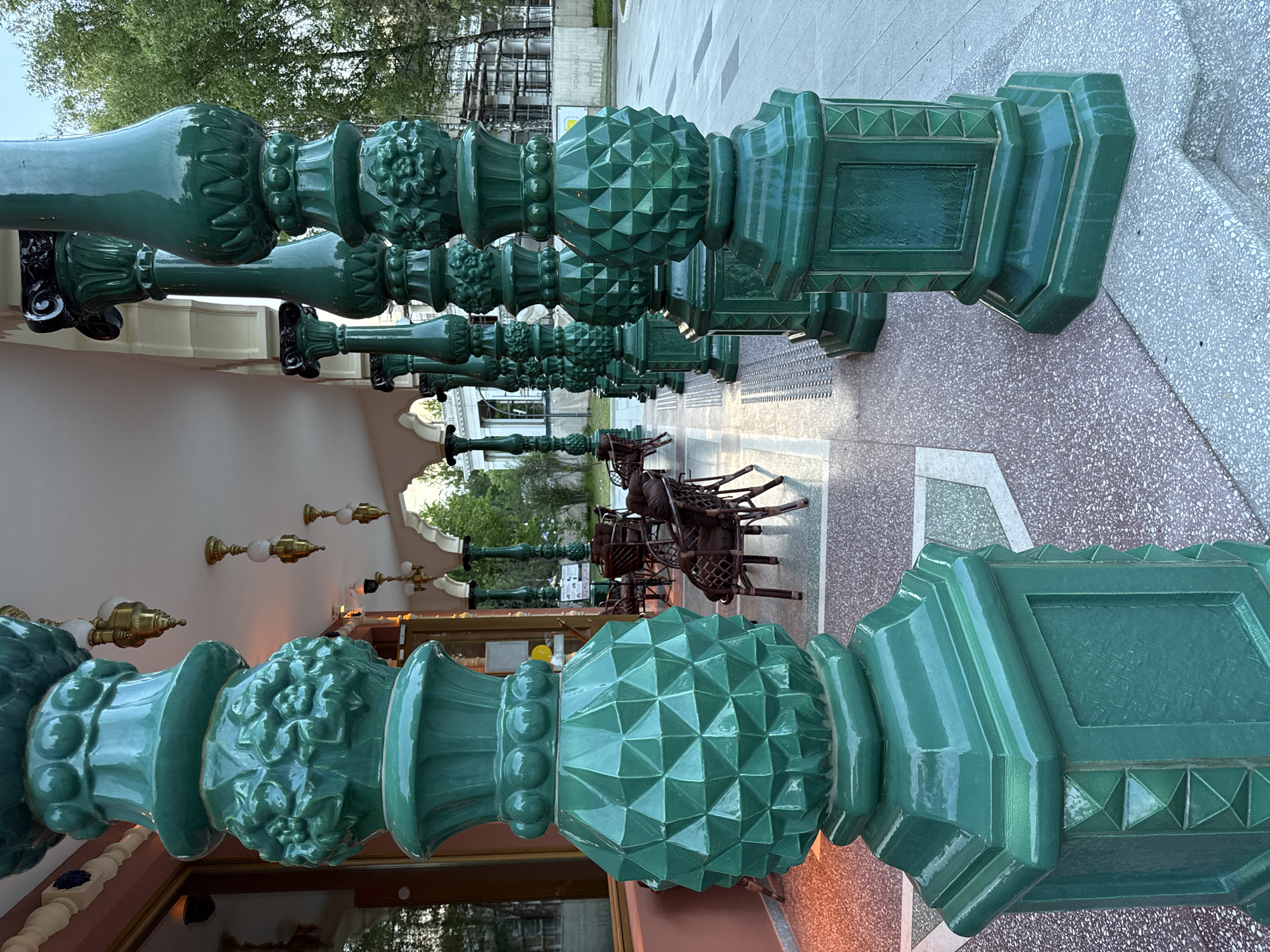 The media file, presented vertically but depicting a horizontally oriented scene, showcases a striking row of ornate, glossy, teal-green balusters or pillars. These decorative elements are made of glazed ceramic or terracotta, exhibiting various intricate patterns including raised floral motifs, spherical embellishments, and distinct geometric, faceted designs resembling pinecones or pyramids. The vibrant green color is consistent across all visible pieces, reflecting light with a subtle sheen.
The pillars line what appears to be an outdoor promenade or walkway, paved with a patterned grey and light-brown speckled surface. To the left (in the original image's orientation), a building facade with arched openings and vintage-style wall-mounted light fixtures can be seen. In the background, beyond the pillars, there are stacked brown wicker chairs, suggesting an outdoor cafe or restaurant area. Further in the distance, lush green trees and a building structure, possibly under construction with scaffolding, are visible.
The scene appears devoid of people, suggesting a quiet moment. The lighting is soft and even, indicating either an overcast day or possibly late afternoon/early evening. There are no harsh shadows. The overall atmosphere is one of serene elegance.
A faint piece of text, "VONA.RU," is visible on a banner associated with the background construction, providing a specific location clue consistent with the given context of Moscow, Russia.