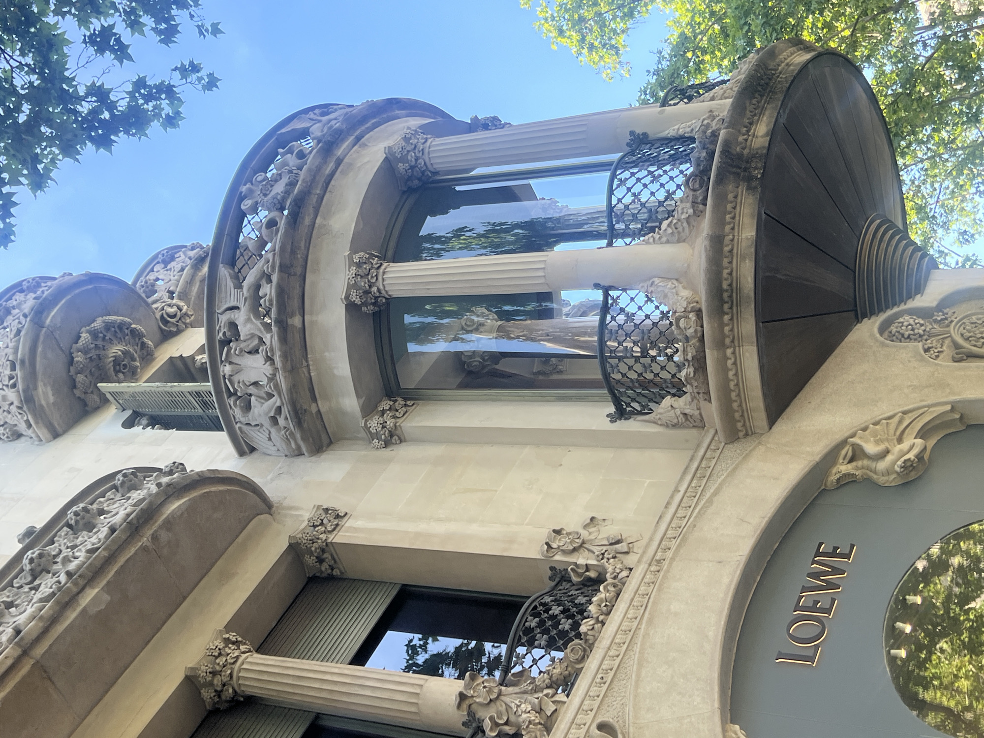 The image shows the facade of a building in Barcelona, Spain, specifically the Casa Batlló, designed by Antoni Gaudí. The architecture is characterized by its organic, flowing shapes and intricate detailing. The building features balconies with wrought-iron railings adorned with whimsical sculptural elements, reminiscent of dragons or mythical creatures. The facade is made of stone, with columns supporting curved balconies and ornate decorative carvings, including floral motifs and stylized figures.
A prominent element in the lower left of the image is the entrance to a store with the "LOEWE" logo displayed in gold lettering. The store has a large arched display window, partially obscured by reflections of trees. The upper portion of the building shows curved windows, some with shutters, and more decorative stonework. Green trees with lush foliage frame the building, contrasting with the blue sky. The overall impression is one of artistic and architectural splendor, typical of Gaudí's work.