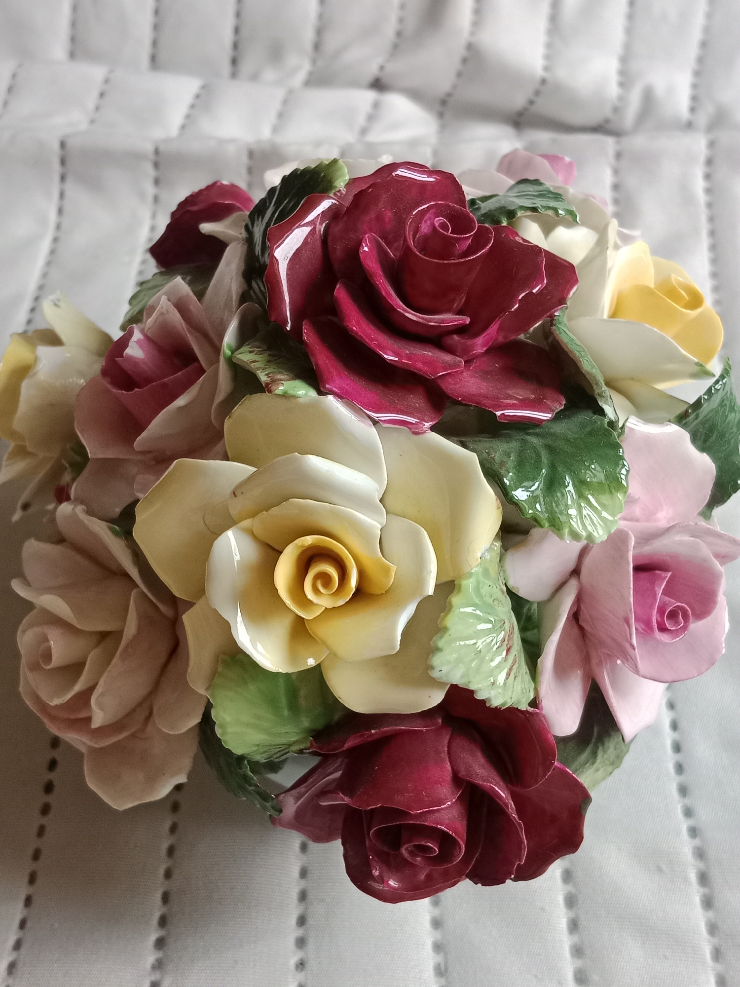 The image shows a close-up, overhead view of a ceramic rose bouquet. The bouquet consists of numerous roses in various shades of pink, red, and yellow, interspersed with green leaves. The roses are intricately detailed with delicate petals and curled centers. The ceramic piece is resting on a white, textured surface that appears to be a quilted fabric, possibly a tablecloth or bedspread. There are no people visible in the image. The scene is static and purely an arrangement of decorative objects. There is no information provided in the image or its context to suggest a location in Bydgoszcz, Poland.