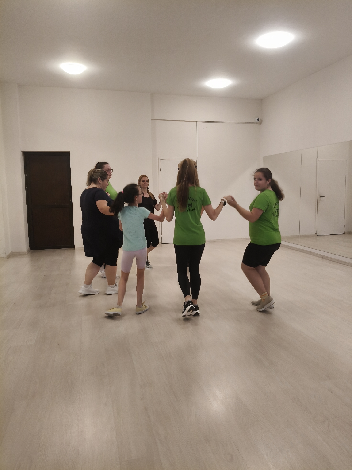 This media file captures six females, ranging from a young child to adults, engaged in what appears to be a dance or movement class. They are positioned in a loose circle, holding hands. Three individuals are wearing matching lime green t-shirts with a logo on the back, black leggings or shorts, and athletic shoes. One adult wears a black top and black capris, another is in a black dress/tunic and black leggings, and a young girl wears a light blue t-shirt, light pink/purple shorts, and yellow sneakers.
The scene is an indoor, brightly lit room with a light-colored wooden floor and white walls, characteristic of a dance studio or rehearsal space. Several circular flush-mount ceiling lights illuminate the room. A large mirrored wall on the right reflects a white door and parts of the interior. A dark brown door is visible on the left wall. A small security camera is mounted high on the right wall near the ceiling.
The participants appear focused on their activity, with some looking towards the camera. The matching green t-shirts suggest they belong to the same group or team. The text on the back of the green shirts is a logo featuring two stylized figures, with partially visible words such as "Little" and possibly "Kiyana." The overall impression is one of active engagement in a dance or exercise session. The context places this scene in Plovdiv, Bulgaria.