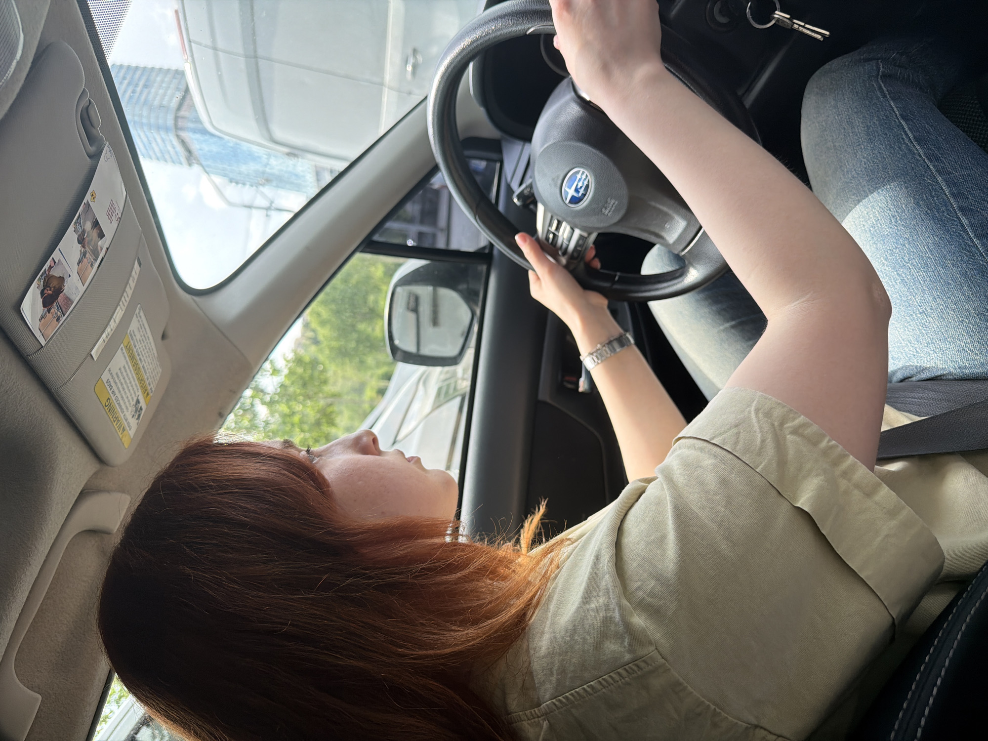 A woman with red hair is sitting in the driver's seat of a Subaru vehicle. She is holding the steering wheel with both hands. The car keys are in the ignition. The woman is wearing a light-colored shirt, jeans, and a watch. The car appears to be stopped, and outside the window, trees and buildings are visible, suggesting an urban setting. The exact location within Tbilisi, Georgia, cannot be determined from the image.