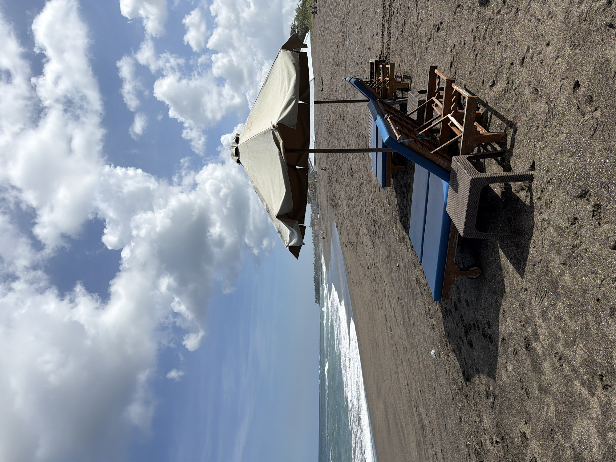 The image depicts a sandy beach in Mengwi, Indonesia, under a partly cloudy sky. The sand is dark, likely volcanic, and shows numerous footprints. In the foreground, two lounge chairs with blue cushions are positioned on the sand, each with a small, brown plastic table next to it. Behind the first lounge chair, another lounge chair is visible. A large, light-colored umbrella provides shade over the chairs. In the middle ground, the ocean stretches out, with waves gently rolling onto the shore. A few people are visible in the distance along the shoreline and in the water. The background shows a line of palm trees and buildings, suggesting a coastal resort area. The overall scene conveys a sense of tranquility and a typical day at the beach.