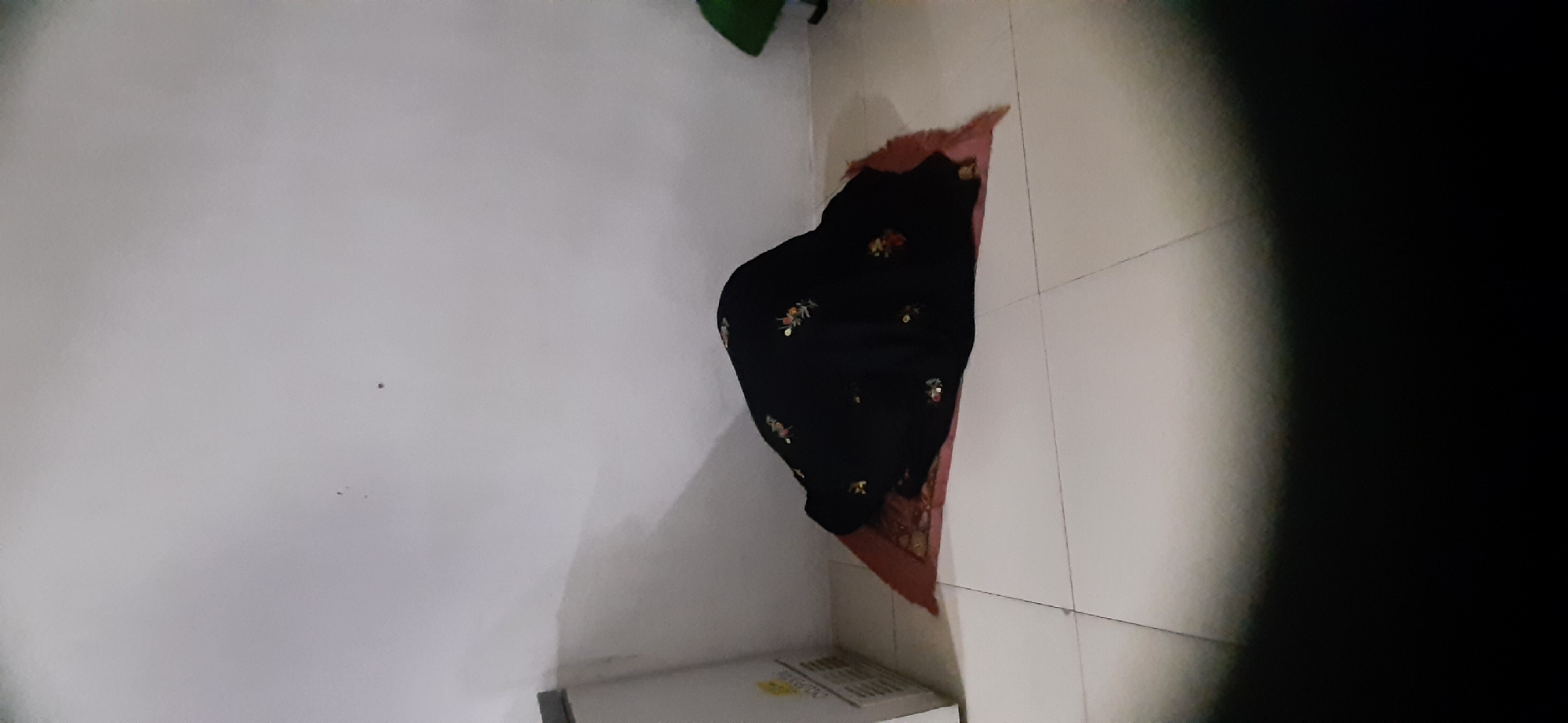A person wearing a black abaya with floral embroidery is performing Salat on a prayer rug. The prayer rug is red with a detailed pattern and is placed on a white tiled floor. The scene is indoors, near a white wall. To the left, a portion of a white refrigerator is visible. The lighting is dim, with a spotlight effect in the foreground. The location is identified as Cisauk, Indonesia.