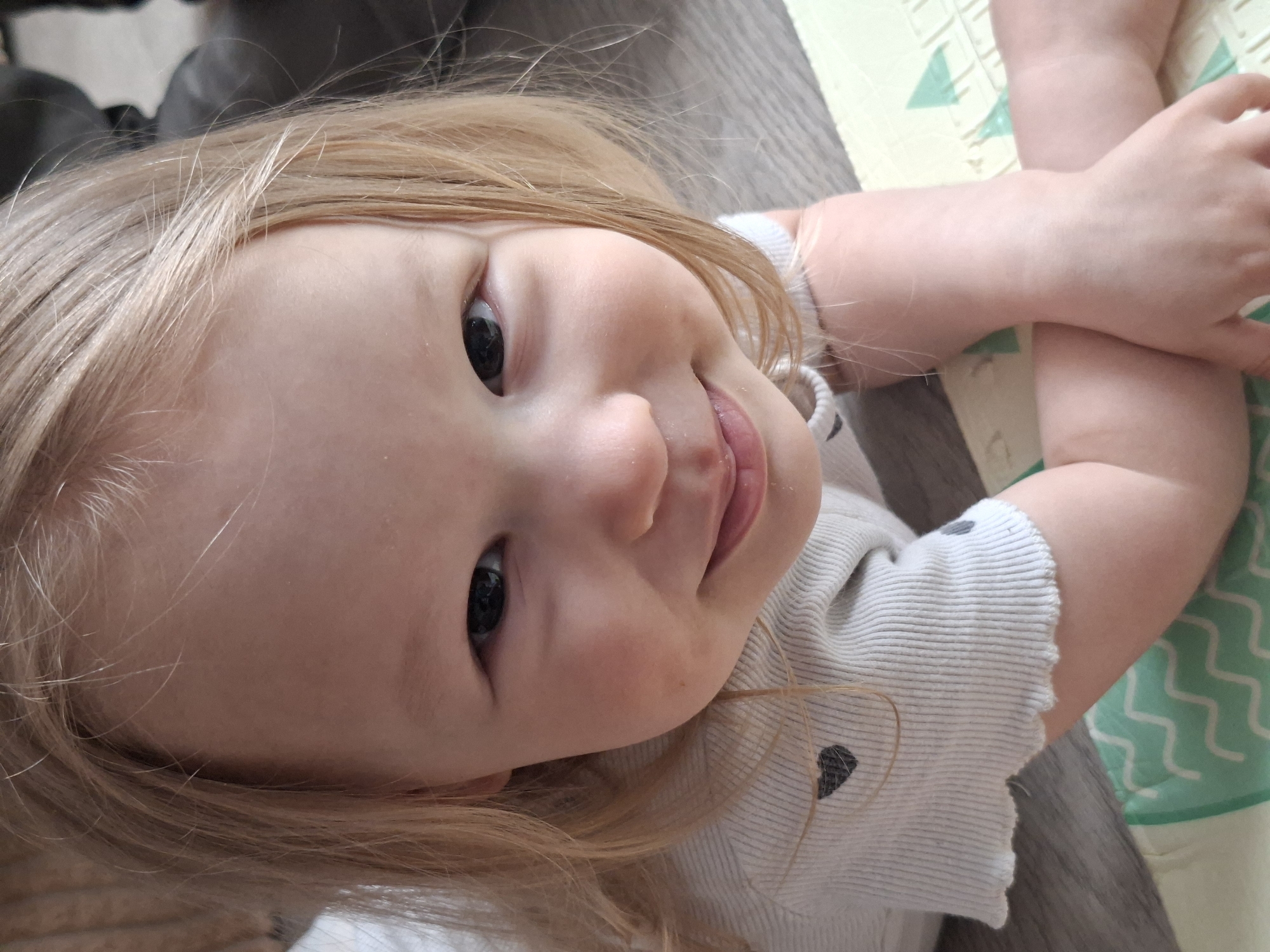 A close-up, slightly tilted, full-face shot depicts a young child with light blonde hair and fair skin. The child gazes upwards towards the camera with dark eyes. Their lips are slightly parted in a subtle smile. They are wearing a light-colored, striped shirt adorned with a small dark heart shape. The child's arms are crossed. The setting includes a gray, wood-like surface on the left and a light-colored play mat with green geometric patterns. The location is Unknown, Unknown.