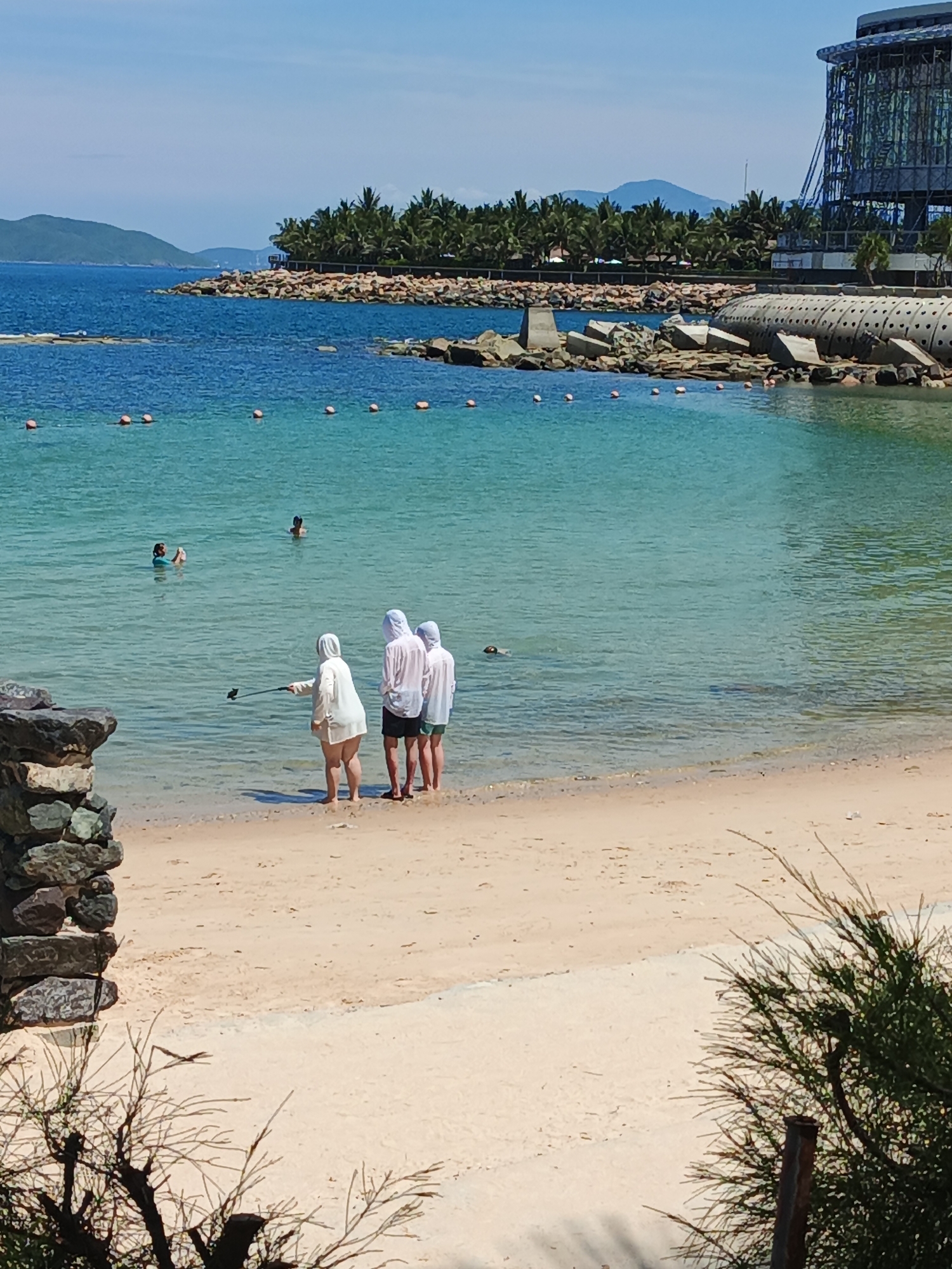 The image captures a serene beach scene with crystal-clear turquoise water and a sandy shore. In the foreground, three individuals stand near the water's edge, one of them holding a selfie stick. The middle person is taking a photo with the selfie stick, while the other two stand behind. Further out in the water, two more individuals are swimming. The background features a coastline with lush green palm trees, rocky breakwaters, and a modern building with a glass facade. The sky is a clear blue, suggesting a sunny day.