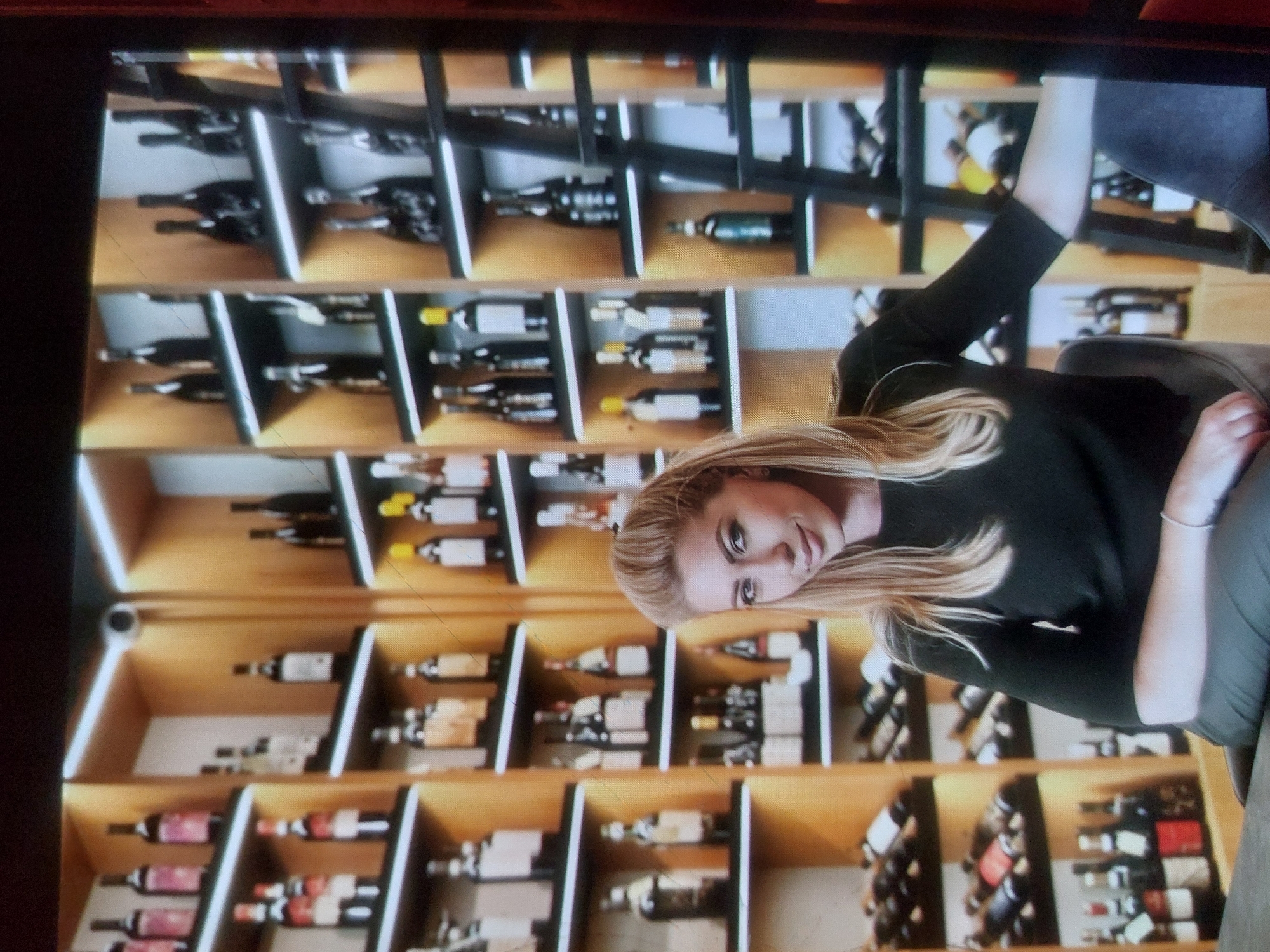 A woman with blonde hair, wearing a black long-sleeve shirt and grey pants, is seated in front of a large wine rack. The wine rack consists of multiple shelves displaying numerous bottles of wine. The location is likely a wine cellar or wine shop, though the exact location in Tsarevo, Bulgaria, is not determinable from the image.