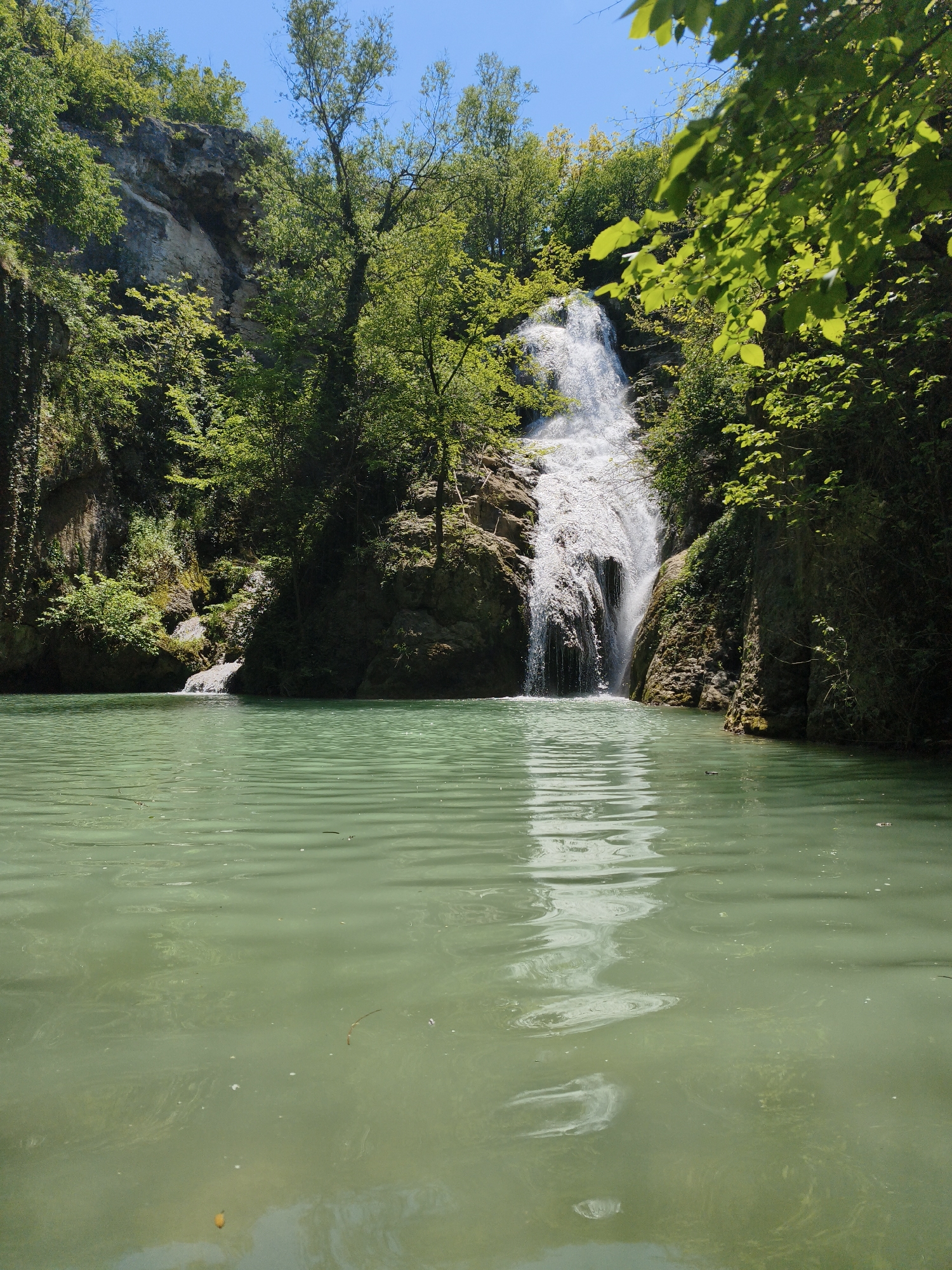 The image depicts the Krushuna Waterfalls, located near the village of Krushuna, north of Letnitsa and east of Hotnitsa, Bulgaria. A cascade of water pours from a cliff face, forming a pool of green water at its base. Lush vegetation covers the surrounding rock formations. The sky is blue and cloudless. Ripples disturb the surface of the water.