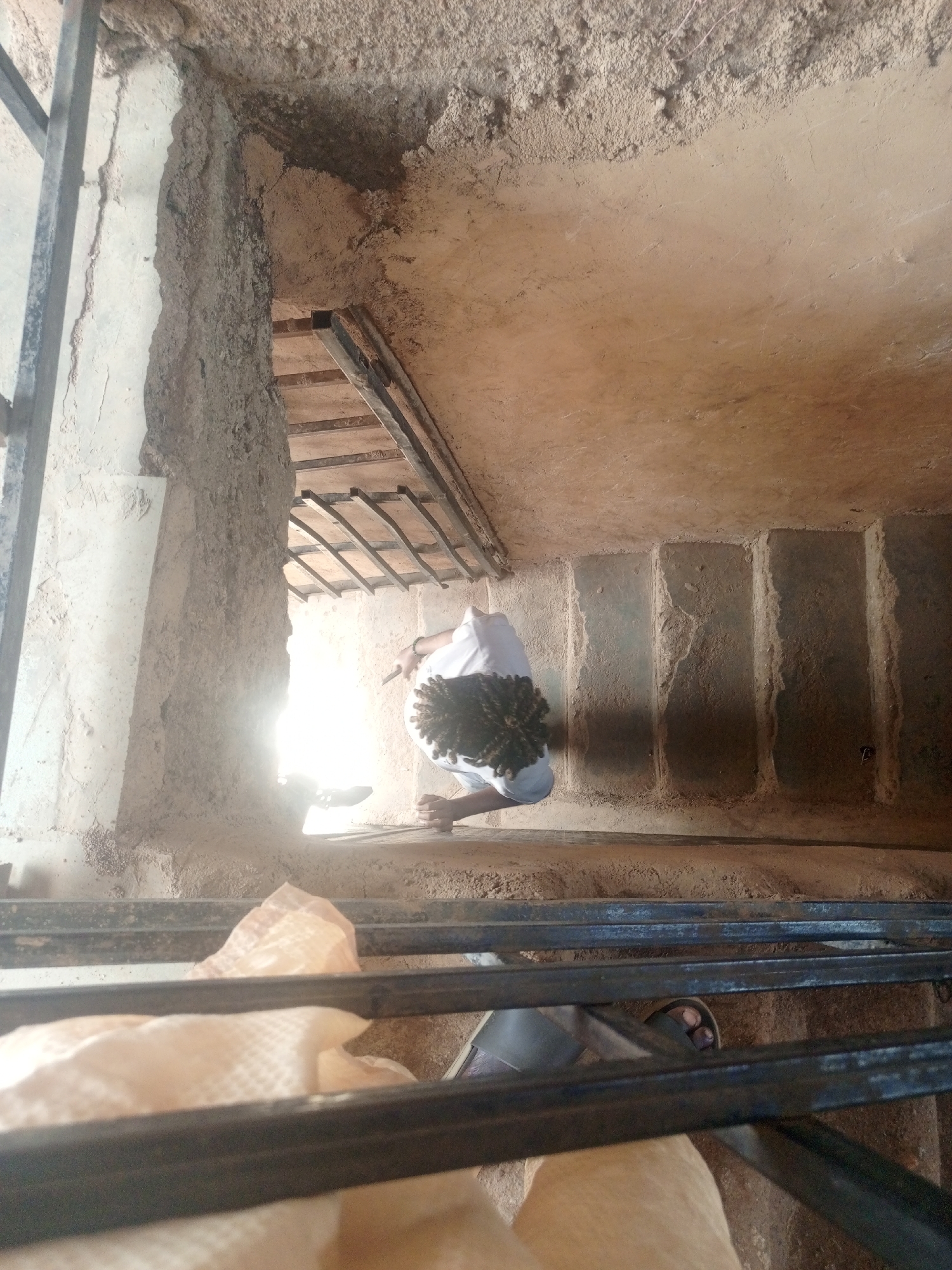 A person is descending a rough, dirt staircase in what appears to be an ancient structure. The person is wearing a white shirt and has dark, curly hair. They are holding a dark object in their right hand. The walls are made of compacted earth and stone, with metal bars visible on the left and in the foreground. The staircase is narrow and the steps are uneven. The lighting is dim, with a bright light source at the bottom of the stairs. The environment suggests an historical or archaeological site. The scene is located in Bukuru, Nigeria.