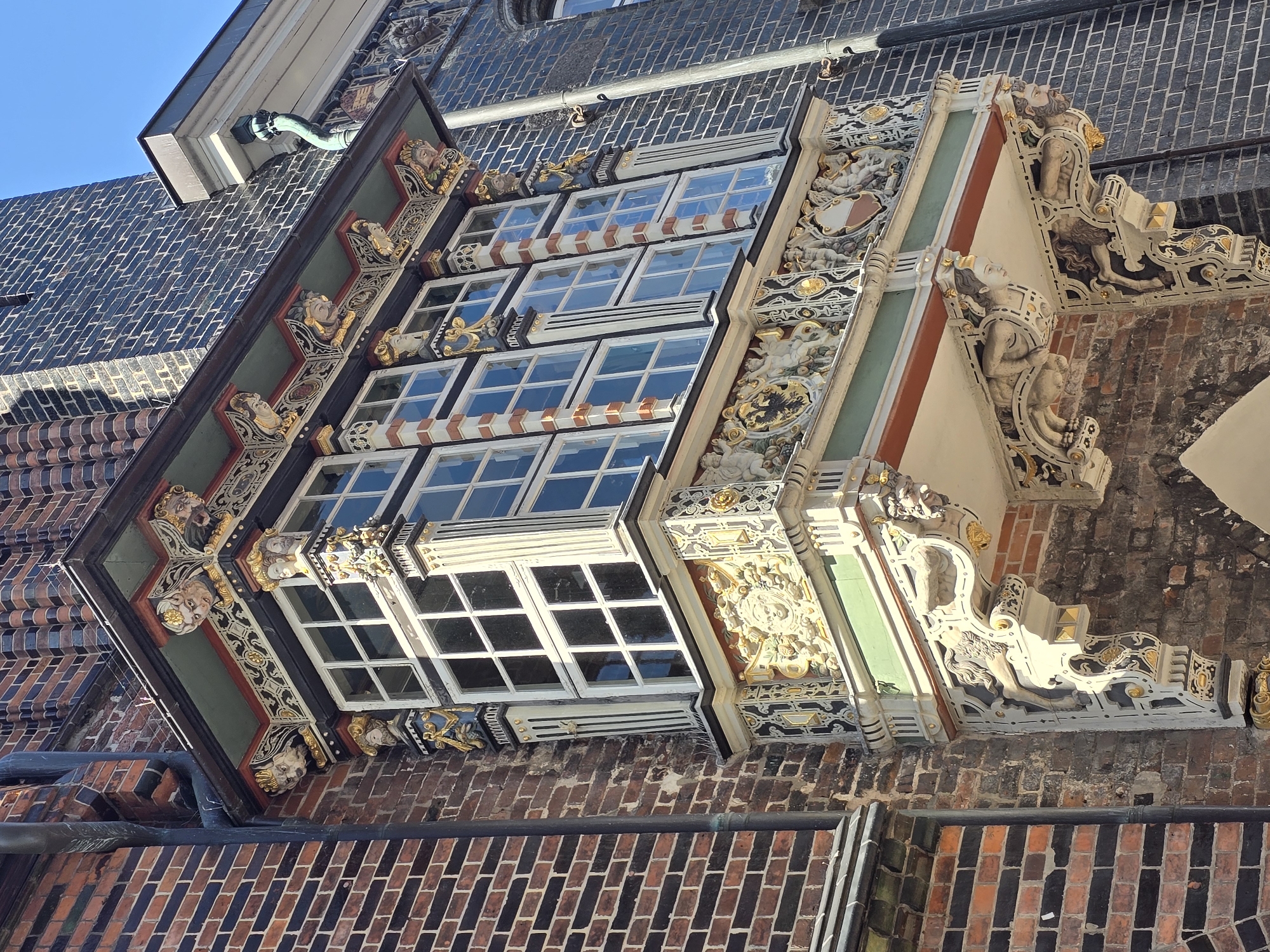 The image shows a detailed view of the "Schabbelhaus" in Lübeck, Germany, a historic merchant house notable for its ornate Renaissance facade. The focus is on a richly decorated bay window, which projects from the brick building. The bay window is supported by elaborate corbels carved into grotesque figures and mythical creatures, with gold accents. The window itself is large, with multiple panes divided by white mullions and transoms, and framed by decorative brickwork and carved elements. Above the window, the eaves are adorned with more grotesque faces and intricate golden carvings. The surrounding walls are constructed of alternating dark and red bricks, characteristic of Lübeck's architecture. A downspout runs vertically along the dark brick wall on the right. The sky above is clear and blue.