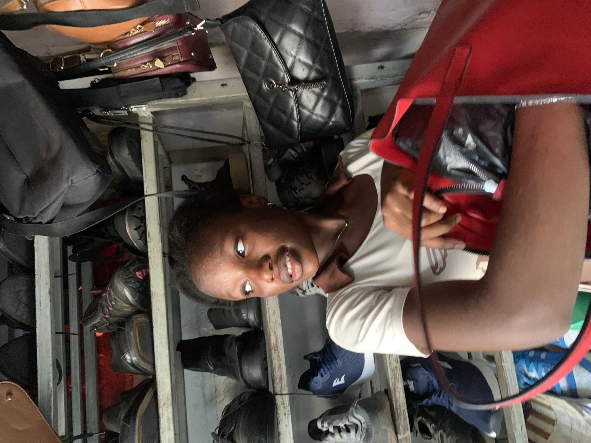 A young Black woman stands in front of shelves filled with shoes and bags, possibly in a market stall in Abuja, Nigeria. She is wearing a light-colored t-shirt with a brown collar and is looking to her right with her mouth slightly open. Her right arm is holding open a large red bag, revealing a black item inside with a zipper. The shelves are filled with various types of footwear, including boots, sneakers, and dress shoes, as well as several handbags of different colors and styles, including a black quilted handbag with a tassel. The background appears to be a wall. The lighting suggests an indoor or covered outdoor space.