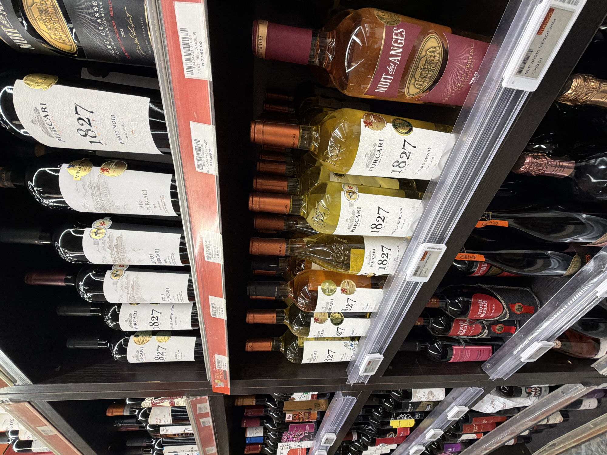 The image depicts a shelf in a retail store in Igboefon, Nigeria, stocked with various bottles of wine. The shelf is made of dark brown wood. A range of wines, primarily red and white, are displayed, including brands like Purcari and Nuit des Anges. The prices of the wines are indicated on shelf tags.