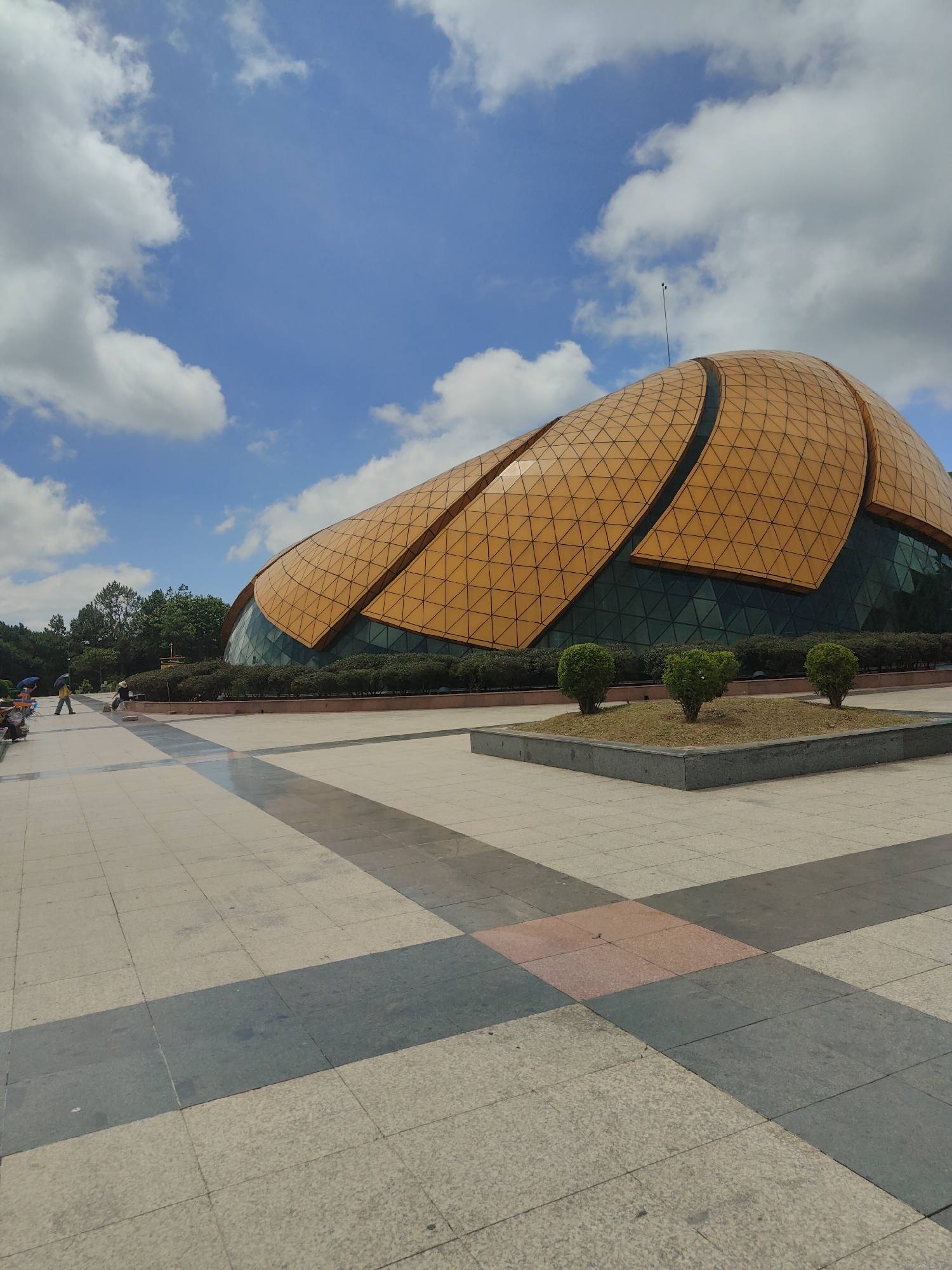 This image captures the Da Lat Flower Garden Exhibition Center in Da Lat, Vietnam, under a partly cloudy blue sky. The prominent structure is a large, curvilinear building with a golden-orange segmented roof, featuring a geometric triangular grid pattern. The lower section of the building consists of a dark, reflective glass facade. A slender, dark antenna-like mast extends from the highest point of the roof.
In the immediate foreground, a wide paved plaza extends across the bottom half of the image. The paving consists of large square and rectangular tiles in varying shades of light grey, dark grey, and some reddish-brown sections, forming a linear and grid-like pattern. Several raised, dark-grey stone planters are spaced across the plaza, each containing three small, neatly trimmed green spherical bushes.
Towards the left side of the plaza, a few individuals are visible in the distance. One person is holding a blue umbrella, another a yellow umbrella, and a third appears to be pushing a small cart or trolley. Beyond the paved area, a line of dense green shrubs borders the building's base, followed by a thick band of mature trees with lush green foliage, forming a natural backdrop. The sky is bright blue with scattered, fluffy white cumulus clouds. The scene is well-lit by natural daylight, indicating clear weather.