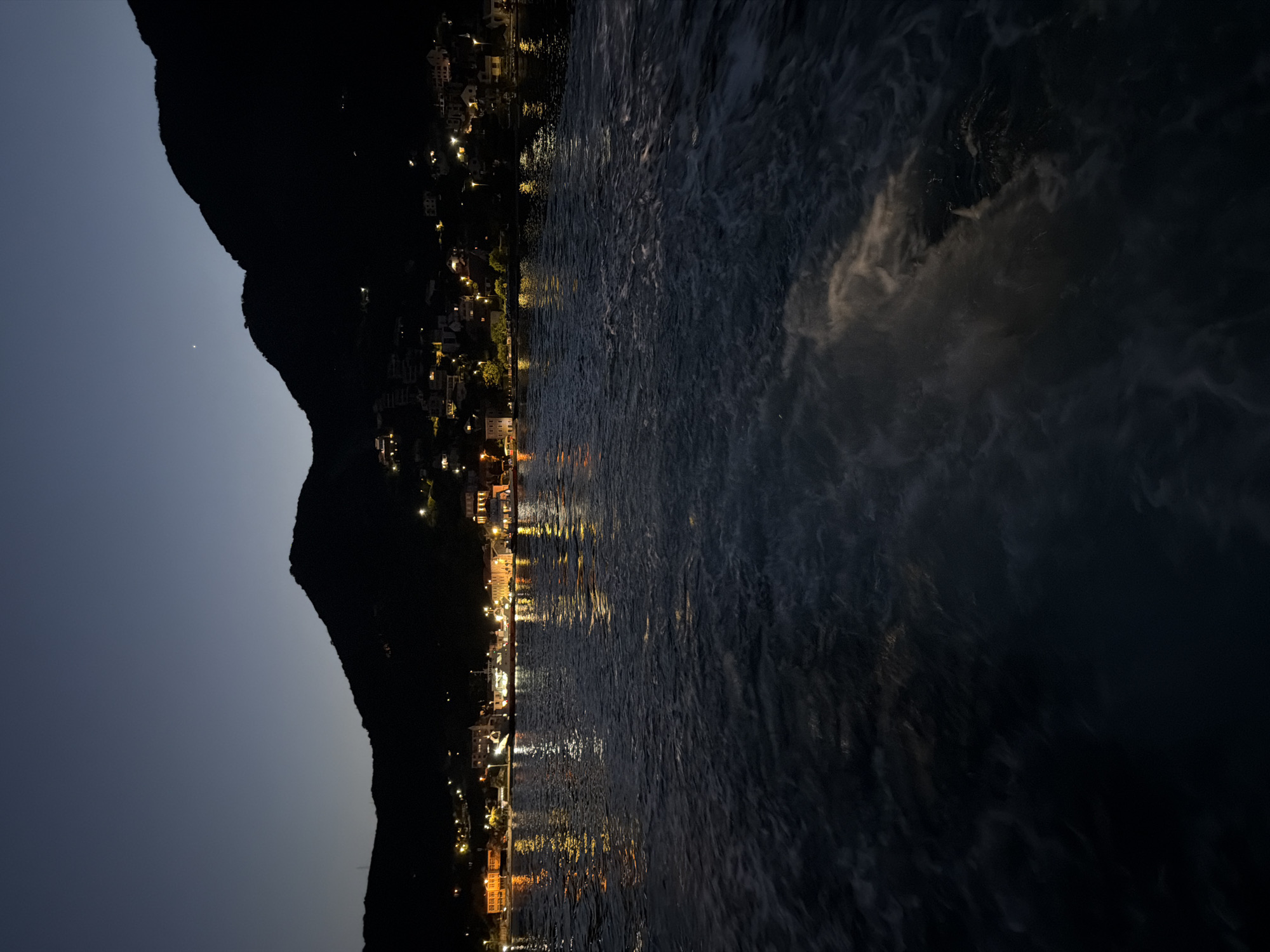 A nighttime long shot captures a body of water in the foreground, with foamy white wakes churning in the lower right quadrant. Beyond the water, a town is illuminated by a series of lights, reflecting off the water's surface. The town is nestled at the base of dark, silhouetted mountains, which rise steeply into the dusky sky. A faint planet or star is visible in the upper right corner of the sky. The scene is likely the Bay of Kotor in Montenegro, near the village of Đurići.
