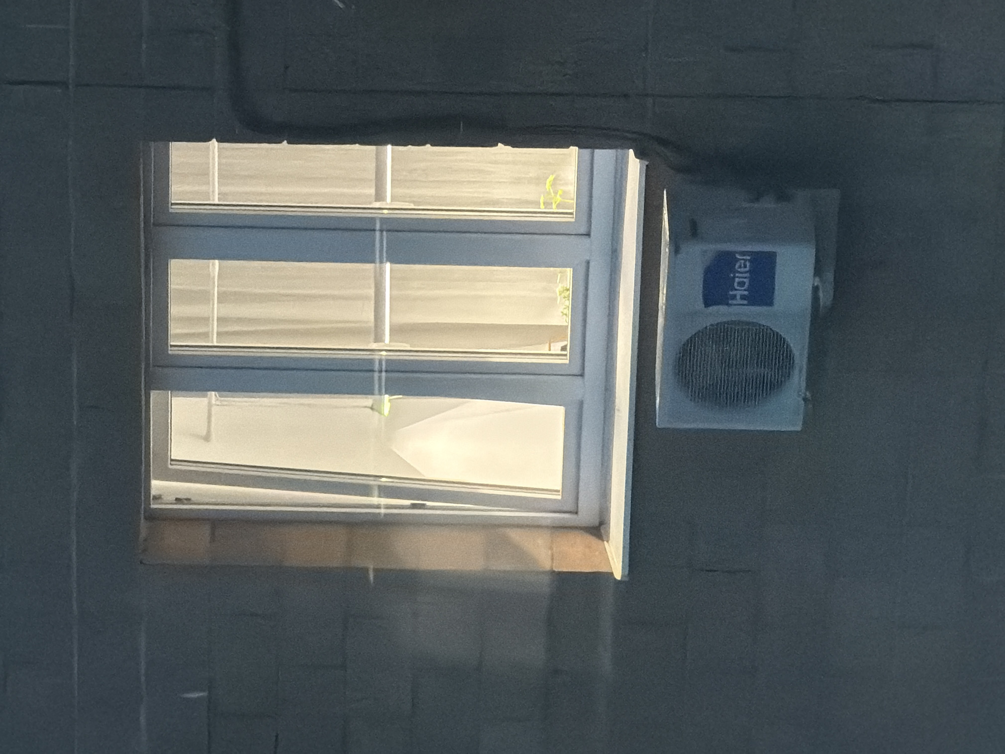 A white-framed window with multiple panes is set into a gray brick wall. The bottom section of the window is slightly ajar. To the right of the window, an air conditioning unit with a "Haier" logo is mounted on the wall. The location is Unknown, Unknown.