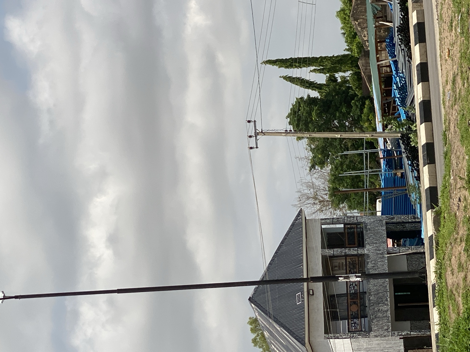 In Jalingo, Nigeria, a two-story building with a grey stone facade and a dark, corrugated metal roof stands prominently. The building features balconies with decorative railings on the upper floor and open ground-level entrances. A tall, dark pole, likely a utility pole, extends vertically through the left side of the frame. In the background, a construction site is visible with stacks of blue pipes and metal scaffolding. Overhead, a power line supported by a wooden pole with ceramic insulators crosses the sky, which is filled with overcast, grey clouds. The scene is bordered by a roadside curb with black and white stripes and a strip of green grass.
