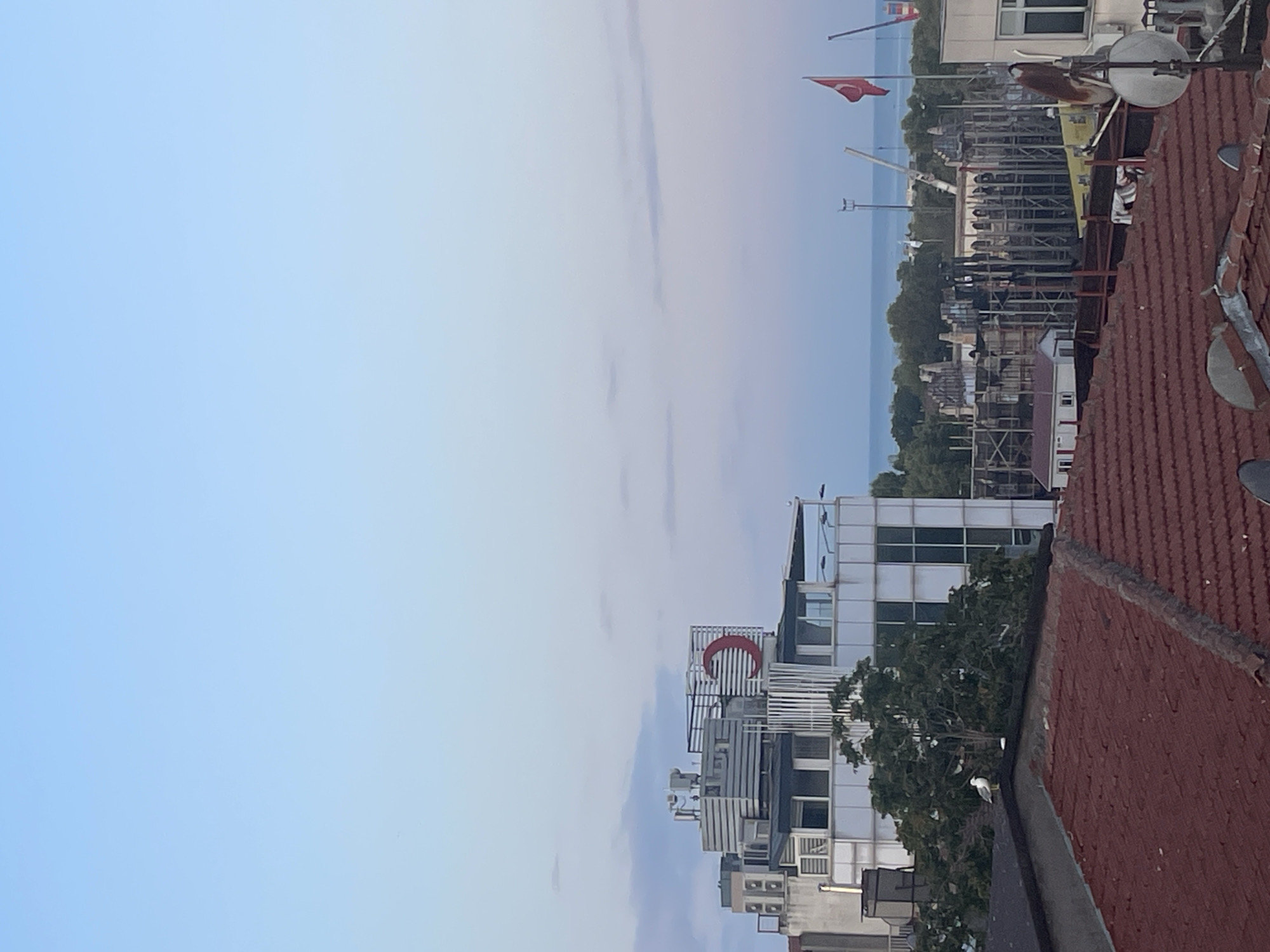 The image shows a view of Kadıköy, Turkey. In the foreground, there are red tiled rooftops. Behind the rooftops, a white building with a large red crescent moon symbol on its facade is visible. Further in the background, more buildings and a Turkish flag are present, with the sea and a hazy horizon in the distance. The sky is a pale blue, indicating either early morning or late evening. A single seagull is perched on the edge of a rooftop in the middle ground. The scene appears to be a cityscape with a coastal element.