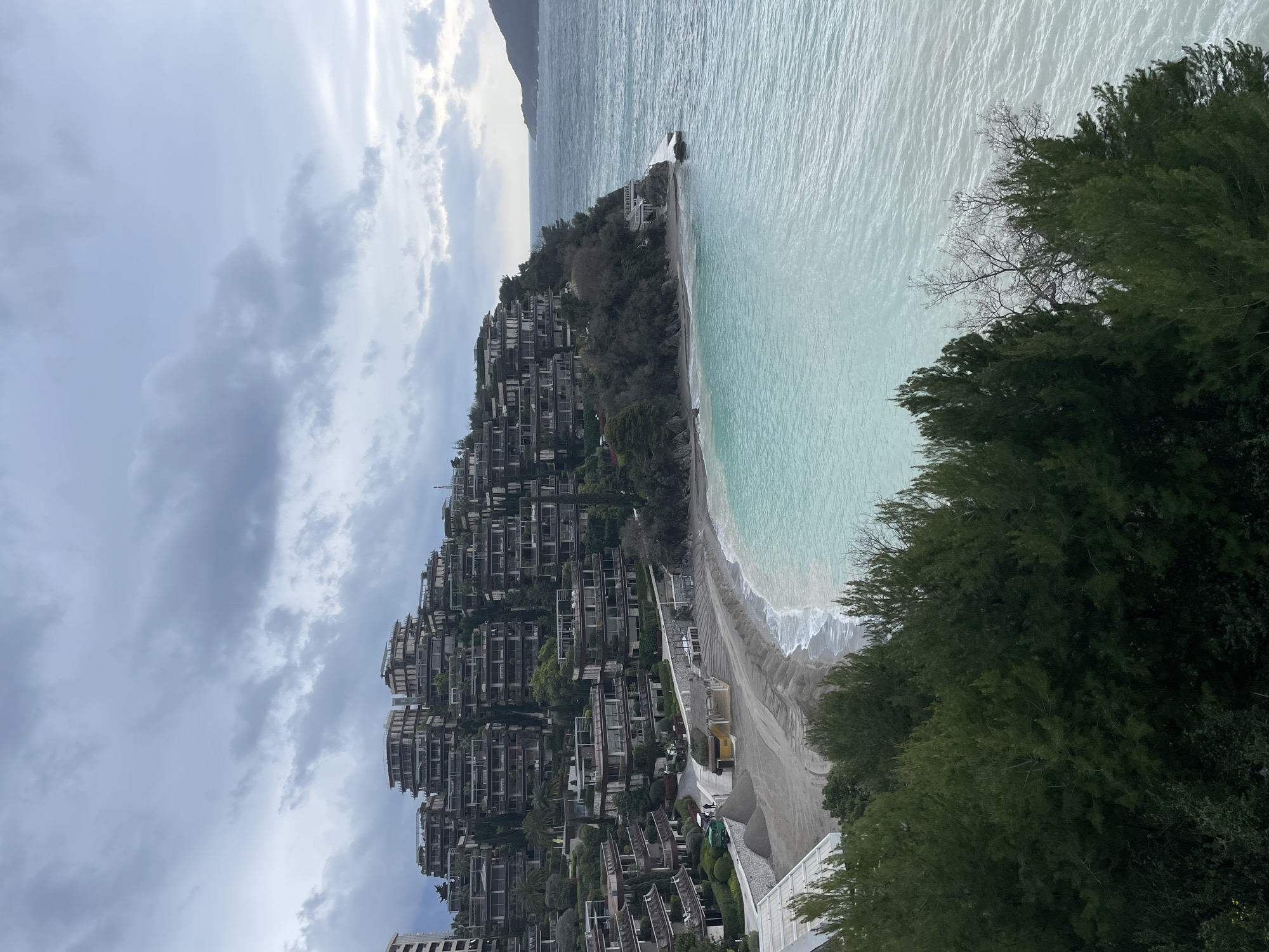 This image depicts a coastal scene on Cape Zavala, Budva, Montenegro, featuring the Dukley Gardens residential complex. The foreground is dominated by dark green coniferous trees. Above these, a crescent-shaped sandy beach, exhibiting piles of light-colored sand, is visible at the base of a steep hillside. Waves break gently on the shore, where the water is a vivid turquoise, transitioning to a darker blue further offshore.
Along the beach, a paved pathway runs parallel to the waterline, separated by a low stone wall. A yellow-roofed structure, likely a beach bar or restaurant, is centrally located on the beach, with white canopy structures positioned to its left. No individuals are visible on the beach or pathway.
The hillside behind the beach is densely populated by a terraced residential complex, consisting of numerous multi-story buildings with flat roofs, extensive balconies, and integrated green spaces including palm trees and other vegetation. These buildings are modern in design, featuring large windows and dark-colored railings. A taller, multi-story building is partially visible on the far left.
To the right, the coastline extends into the sea, where a small white structure with a platform protrudes from the rocky shore. In the distance, a dark landmass, possibly an island or headland, is discernible on the horizon. The sky above is overcast, filled with grey, stratified clouds, suggesting a muted daylight condition. No specific actions or interactions are depicted within the scene.