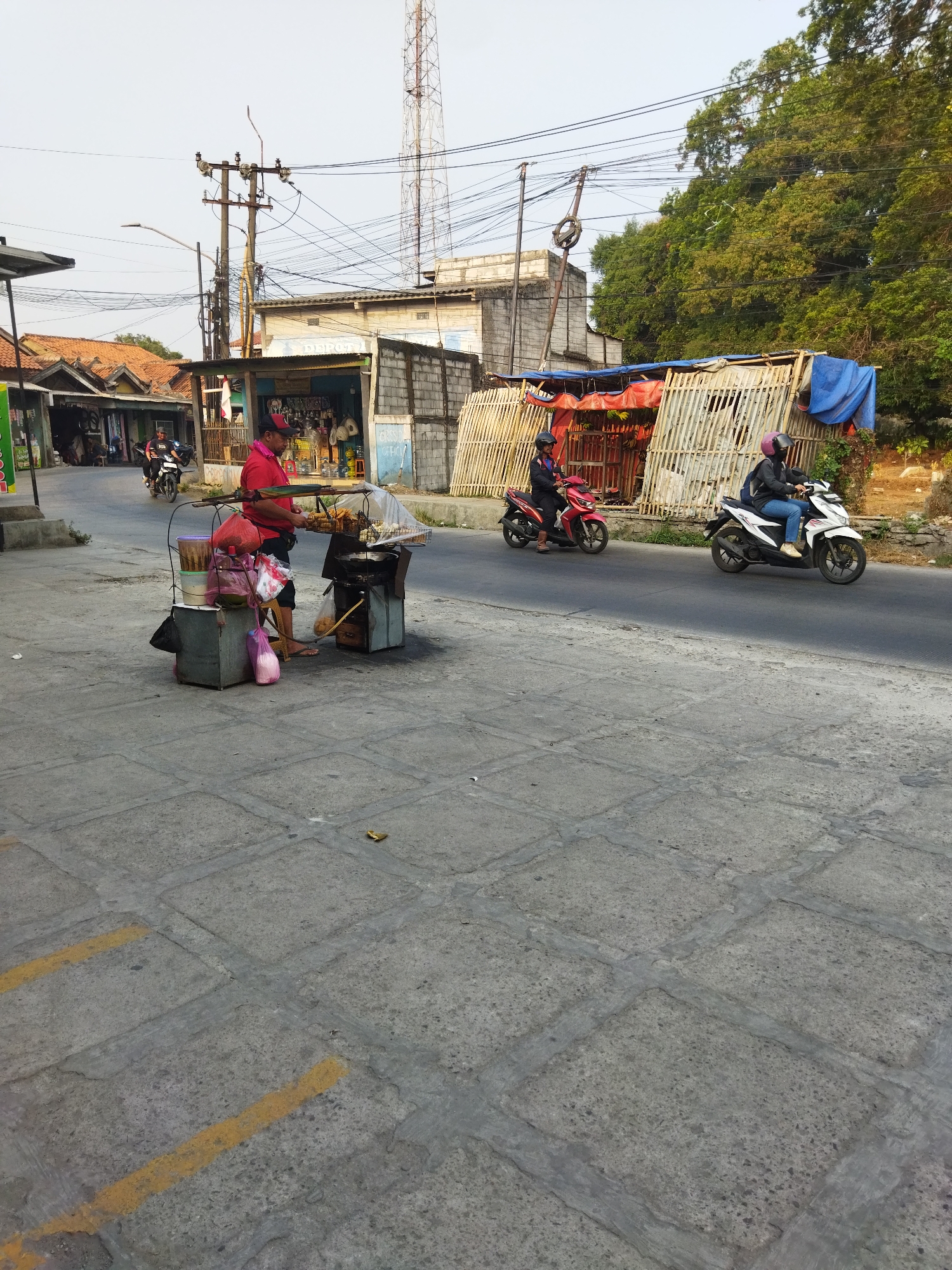 This image captures a street scene in Pisangan Jaya, Indonesia. In the foreground, a street food vendor is preparing food at a cart on the sidewalk. The vendor, a man in a red shirt and black shorts, stands behind a display of food items, some of which are being fried. Adjacent to the cart are several containers and bags, including a pink plastic bag. The sidewalk is paved with large, square concrete slabs, marked with yellow lines.
In the background, a paved road runs alongside the sidewalk. Two motorbikes are traveling on the road; one red motorbike with a rider wearing a dark jacket and helmet, and a white motorbike with a rider wearing a dark jacket, jeans, and a pink helmet. Beyond the road, there are several buildings, including a structure with a corrugated metal roof and a stall with a bamboo-like facade covered partially by a blue tarp. Electrical poles with numerous wires are visible, as is a tall transmission tower in the distance. Green trees form a backdrop on the right side of the image. The lighting suggests it is daytime, possibly late afternoon, with the sun casting shadows.
