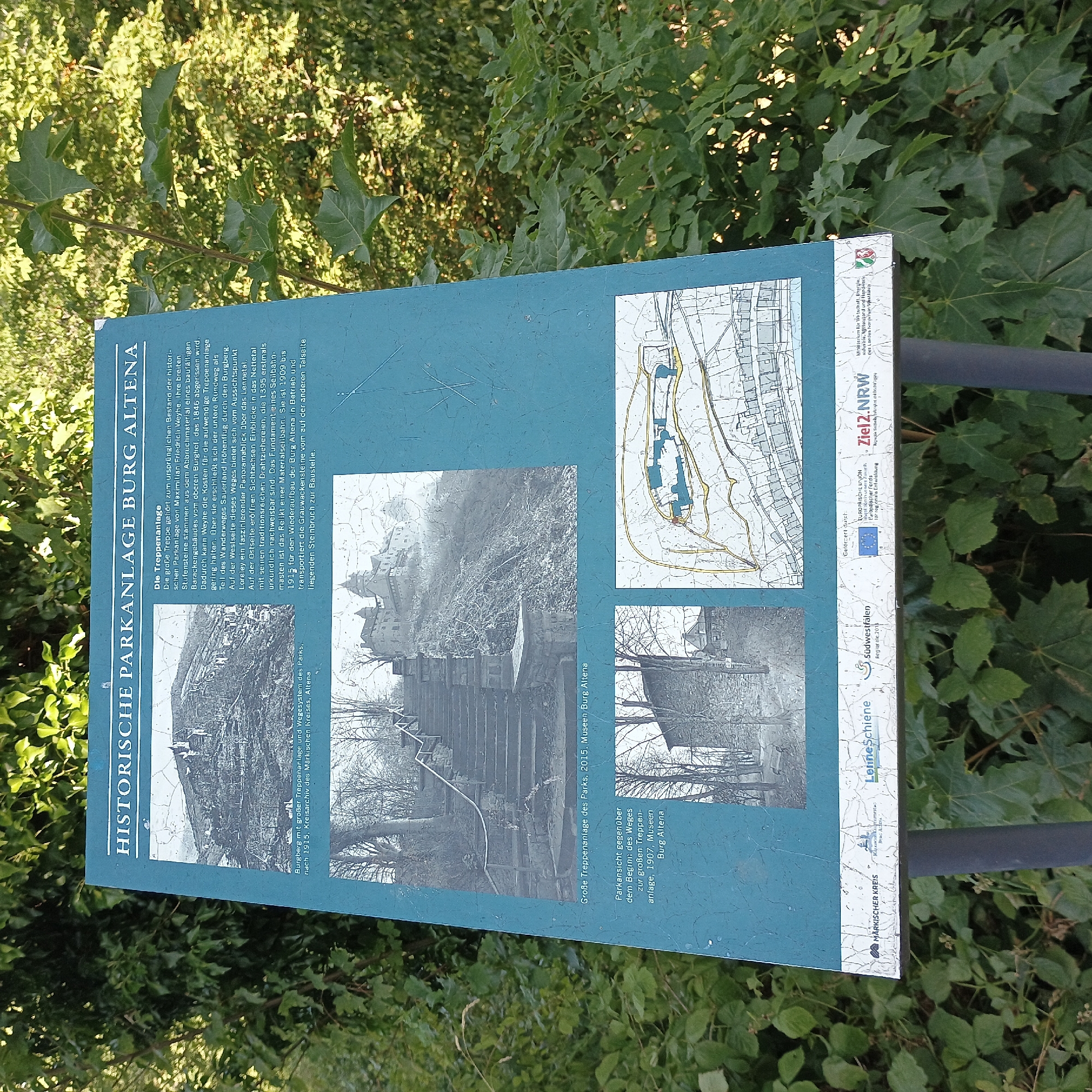 A weathered, informative sign stands in a lush, green outdoor setting, surrounded by dense foliage and trees. The sign itself is a dark teal color and features a white border. It is divided into sections, each containing text and images.
The top section displays a black and white photograph of a sprawling historical park with a prominent castle or fortress situated on a hilltop. Below this photograph is a caption: "Burgberg mit großer Treppenanlage und Wegesystem des Parks, nach 1915, Kreisarchiv des Märkischen Kreises, Altena." This translates to "Castle hill with large staircase system and park's path system, after 1915, District Archive of the Märkischer Kreis, Altena."
The middle section features another black and white photograph showing a grand stone staircase leading up a slope, with trees and the castle visible in the background. This image is captioned: "Große Treppenanlage des Parks, 2015, Museen Burg Altena." This means "Large park staircase, 2015, Museums Burg Altena."
To the right of the staircase photograph, a detailed map is presented. The map outlines pathways and structures, likely depicting the layout of the historical park and its surroundings.
The bottom section of the sign contains several logos and text, indicating funding and official affiliations. These include logos for "Märkischer Kreis," "Südwestfalen," and "Ziel 2.NRW," along with text related to the European Union and governmental bodies.
The overall scene suggests an informational display within the historical grounds of Burg Altena, a castle located in the city of Altena, Germany. The environment is natural and overgrown, with the sign being the central man-made object. No people are visible in the image. The sign is mounted on a dark grey pole.