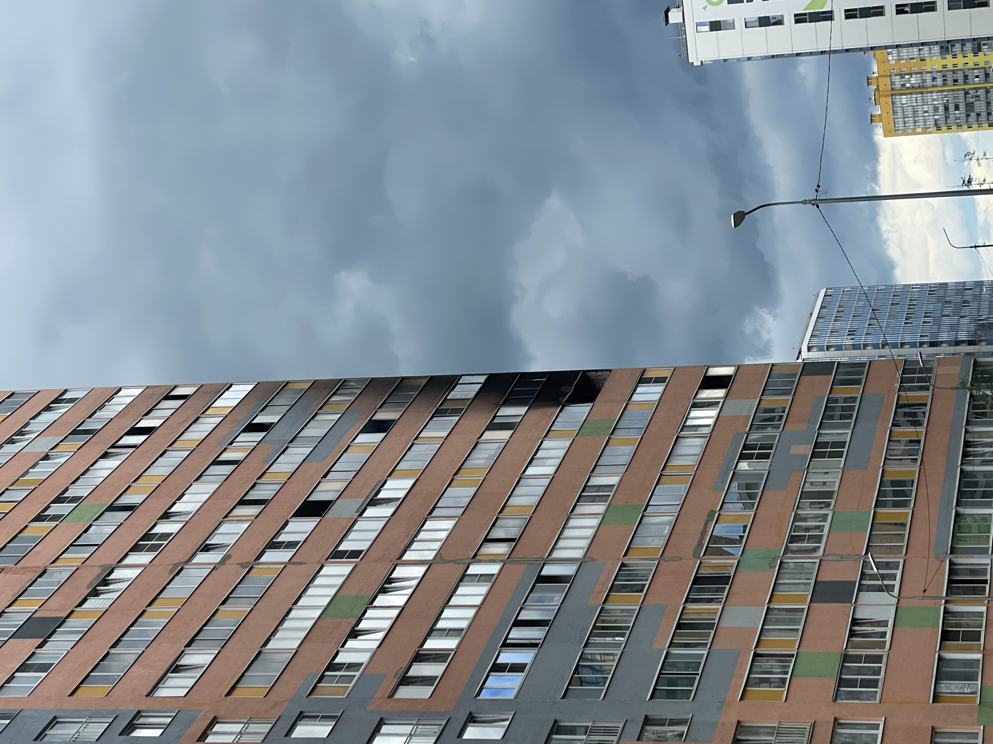 In Murino, Russia, a towering residential building, predominantly salmon-colored with grey and colored accents, is visible on the left side of the frame. The building features numerous windows, many of which are open. A distinct section on the right side of the salmon-colored facade appears scorched and blackened, indicating a fire. To the right of this building, a streetlamp stands with wires extending across the image. Further to the right, two other residential buildings are partially visible: one is a tall, white building with green accents, and another is a yellow and grey building. The sky is overcast with dark, dramatic clouds.