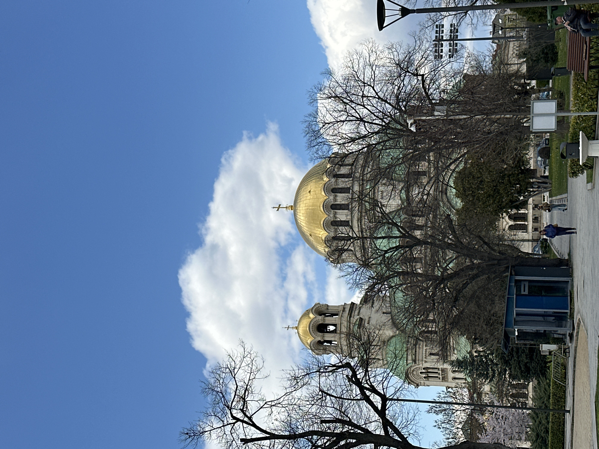 The image depicts the Alexander Nevsky Cathedral in Sofia, Bulgaria, with its distinctive gold domes and intricate architecture. A portion of a city park is visible, with trees, benches, and a few people walking along the pathway. A blue-and-gray building structure is also visible on the right side of the scene. The sky is blue with some clouds. The scene is shot on a sunny day.