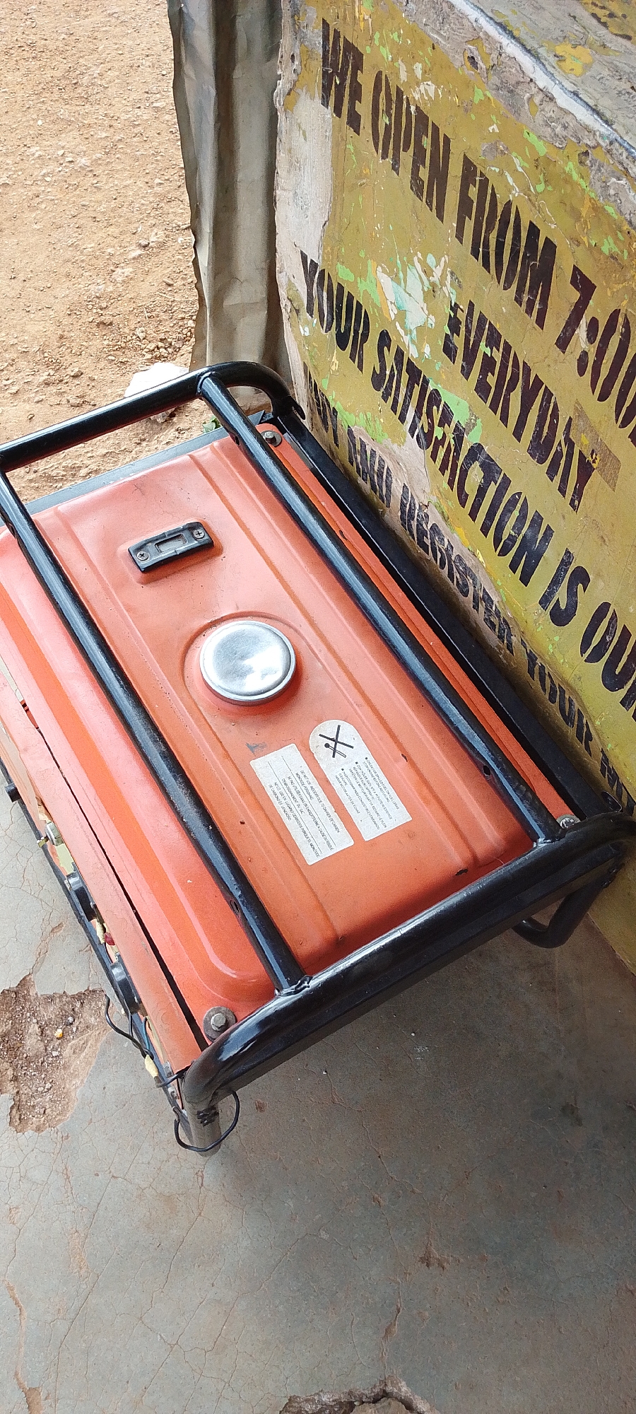 An orange portable generator with a black metal frame is resting on a cracked concrete surface. A faded yellow sign with black lettering is partially visible on the right, reading "WE OPEN FROM 7:00 YOUR SATISFACTION IS OUR". The sign appears weathered and peeling. The ground to the left is sandy and reddish-brown. A portion of a metallic, corrugated structure is also visible behind the generator and sign. There are no people in the image.