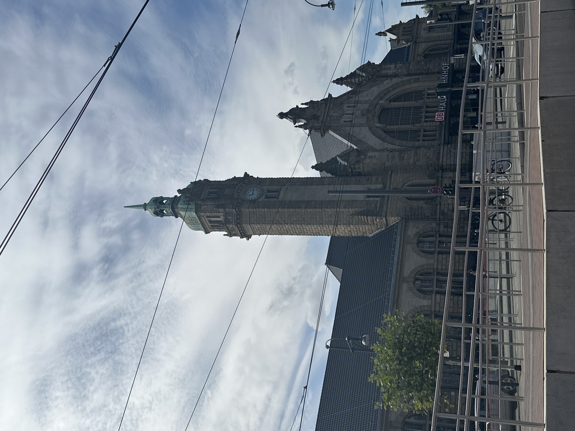 The image shows the Krefeld Hauptbahnhof, a train station in Krefeld, Germany. The building is made of stone with a tall clock tower. The sky is cloudy and there are overhead wires in the foreground, likely for a tram or train. A metal railing runs across the image in front of the building, with a sidewalk and street beyond. A car is visible on the street, and a bicycle is parked nearby. The sign above the entrance reads "DB HAU HNHOF", indicating it is a Deutsche Bahn (German Railways) station.