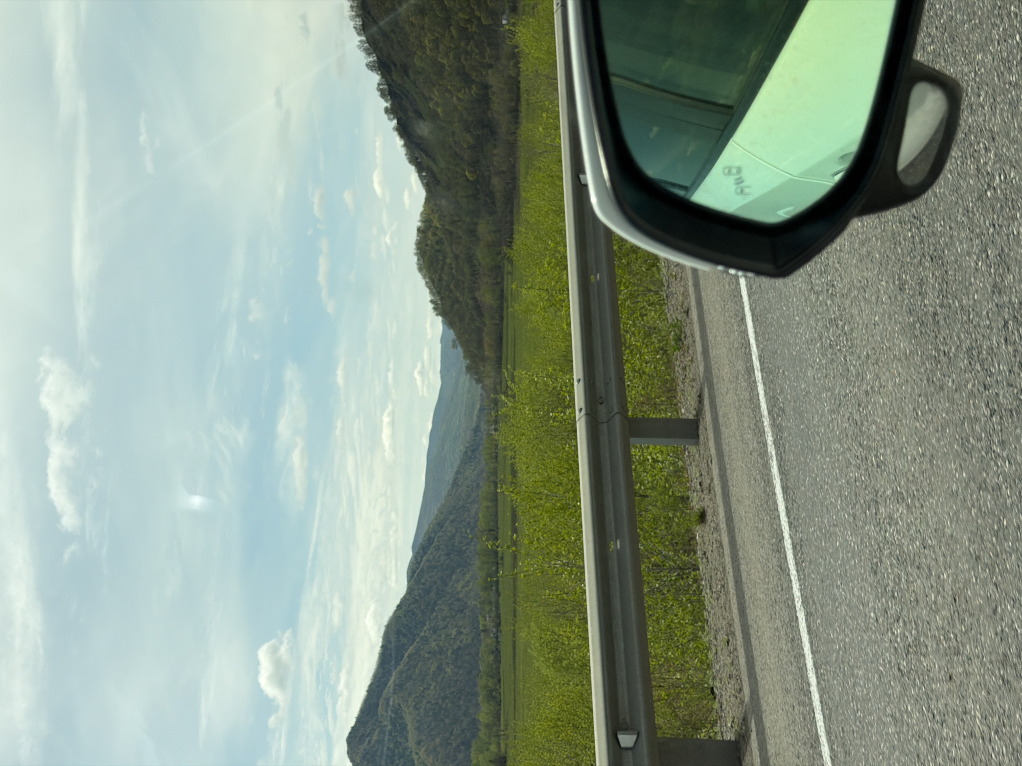 The image shows a view from a car traveling on a road near Лондоко-завод, Russia. A mountain range is visible in the distance, with a field of green vegetation between the road and the mountains. A guardrail runs along the side of the road. The car's side mirror is visible in the upper right corner of the image. The sky is partly cloudy.
