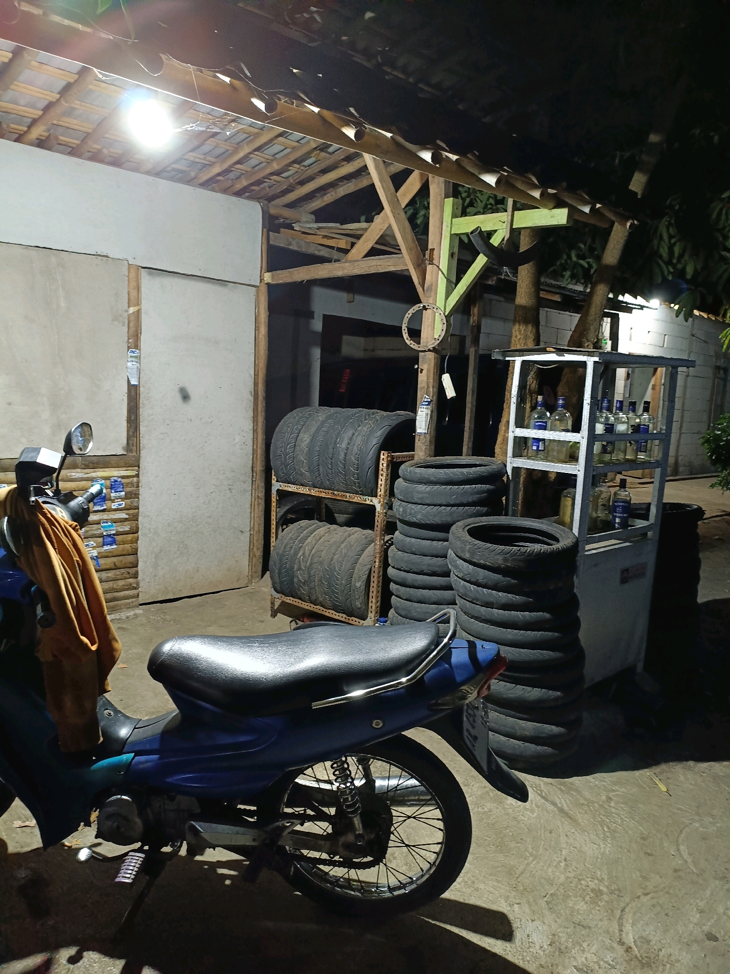The image depicts a nighttime scene in Semanu, Indonesia, likely an outdoor workshop or storage area. In the foreground, a blue motorcycle is parked. Behind the motorcycle, several stacks of tires are visible, with some on metal racks and others piled on the ground. To the right of the tires, a small metal shelf unit displays numerous bottles, appearing to contain liquid. The area is illuminated by a bright overhead light, casting strong shadows. The structure behind the objects suggests a simple, open-air shelter with a wooden roof and possibly concrete or cinder block walls in the background. The ground is paved, and trees are visible in the background, indicating an outdoor setting. There are no people visible in the image.