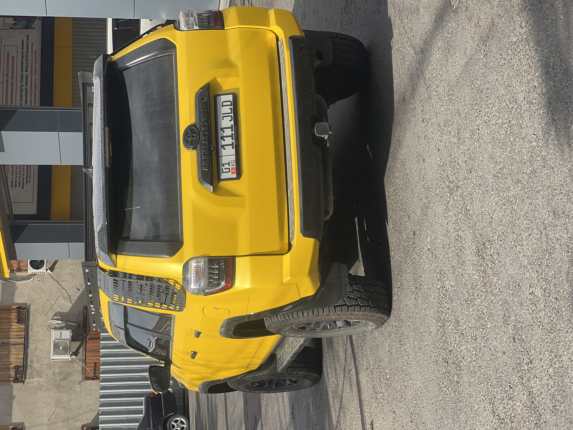 In Bishkek City, Kyrgyzstan, a bright yellow Toyota 4Runner is parked on an asphalt surface. The vehicle has off-road tires and aftermarket accessories, including a roof rack and a cargo cage on the rear side window. The license plate reads "01 111 JLD KG". The vehicle is positioned in front of a building with a gray concrete wall and wooden shutters, and behind a corrugated metal fence. Several air conditioning units are visible mounted on the wall. The scene appears to be in an outdoor urban or industrial area, with shadows indicating sunlight.