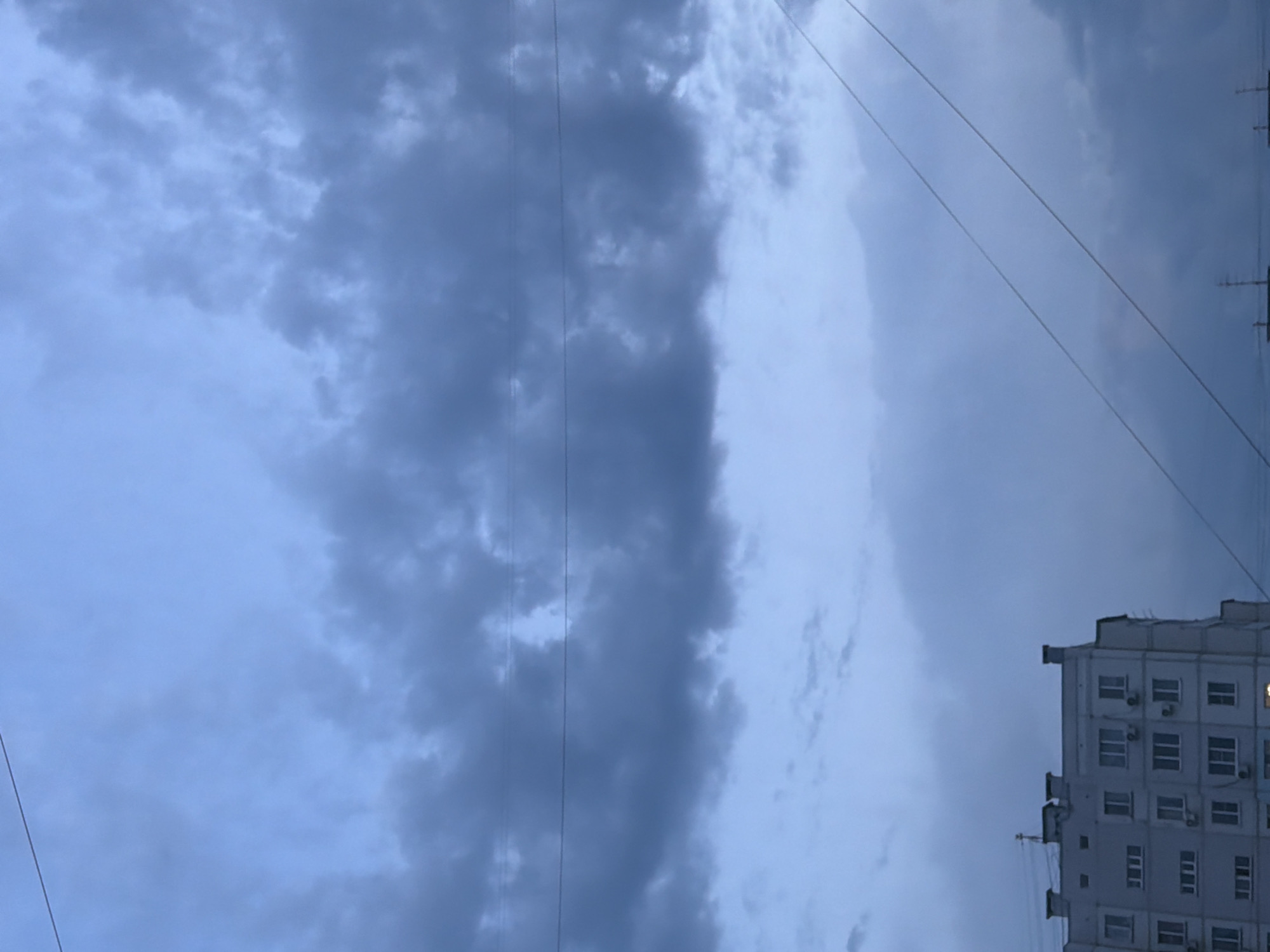 The image shows a cloudy, overcast sky above a residential building and power lines. The building appears to be a multi-story apartment complex with numerous windows and balconies. The sky is a muted blue-gray, with dark clouds dominating the upper portion of the frame. Several thin power lines stretch across the sky, intersecting the building and the clouds. The overall scene suggests a gloomy or pre-storm atmosphere. The location is identified as Khabarovsk, Russia. There are no people or specific actions visible in the image.