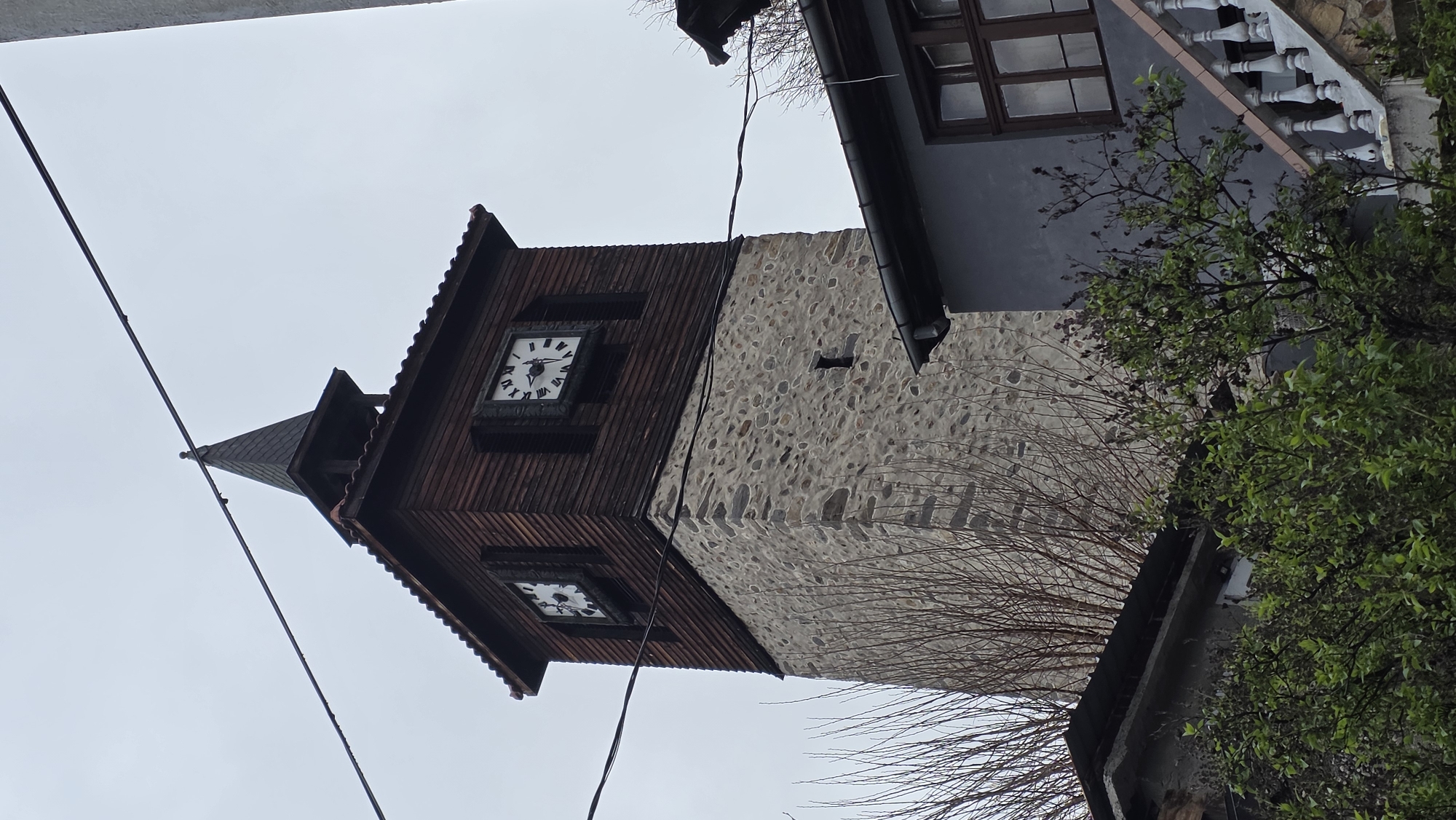 The image depicts the clock tower in the city of Blagoevgrad, Bulgaria. The tower has a stone lower section and a wooden upper section with two clock faces. A small conical roof caps the tower. Power lines cut across the sky in the background. Part of a building with a balcony and windows is visible to the right of the tower, partially obscured by tree branches. The sky is overcast.