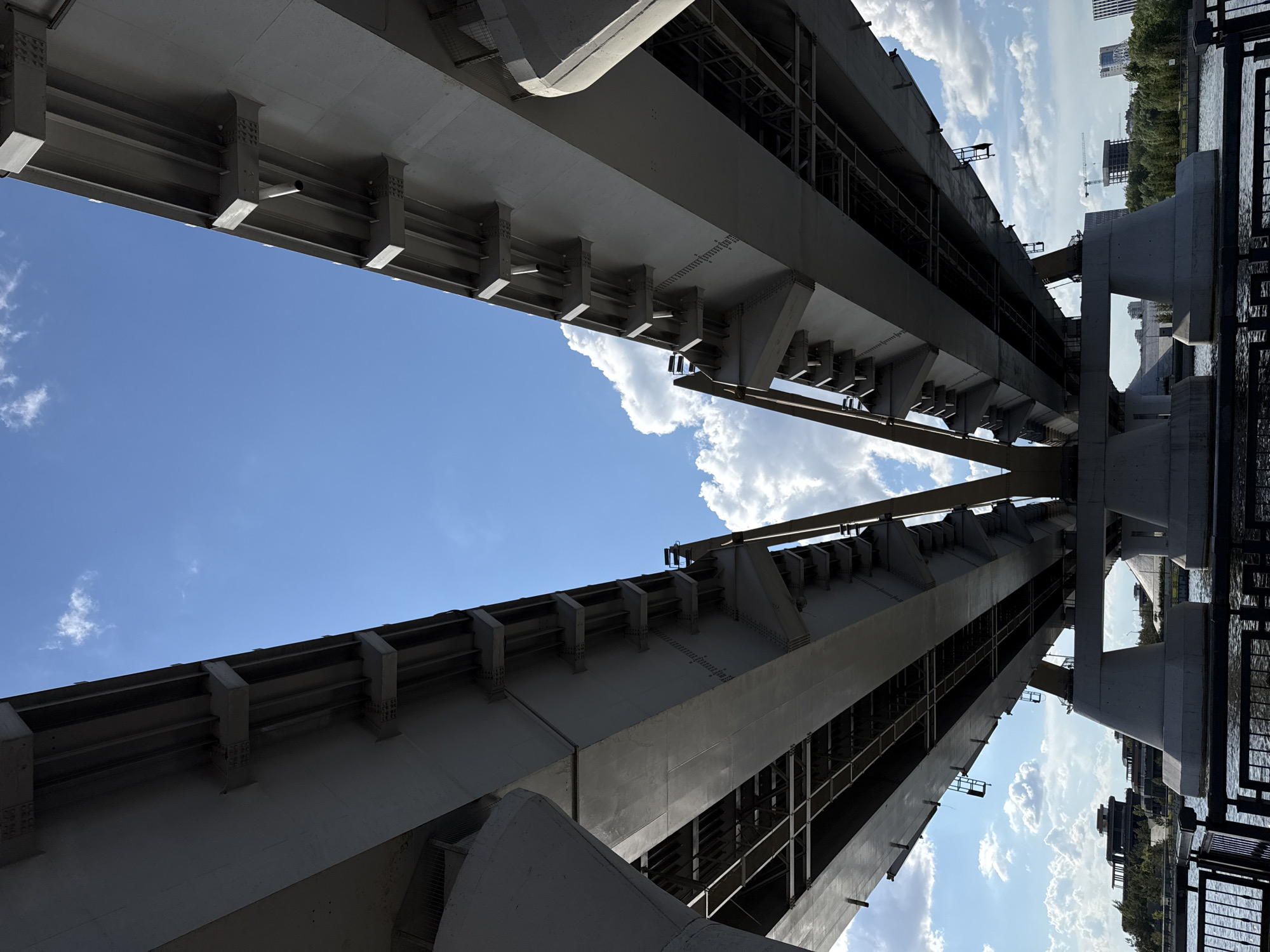 The image shows the Millennium Bridge in Kazan, Russia, viewed from below. The steel and concrete structure of the bridge dominates the frame, angled upwards to showcase its supports and beams against a backdrop of blue sky with scattered white clouds. The bridge spans over water, with the opposite bank visible on the right, including buildings and trees. The viewpoint is low, emphasizing the scale and engineering of the bridge. Reflections of the sky and surrounding structures can be seen on the water's surface.