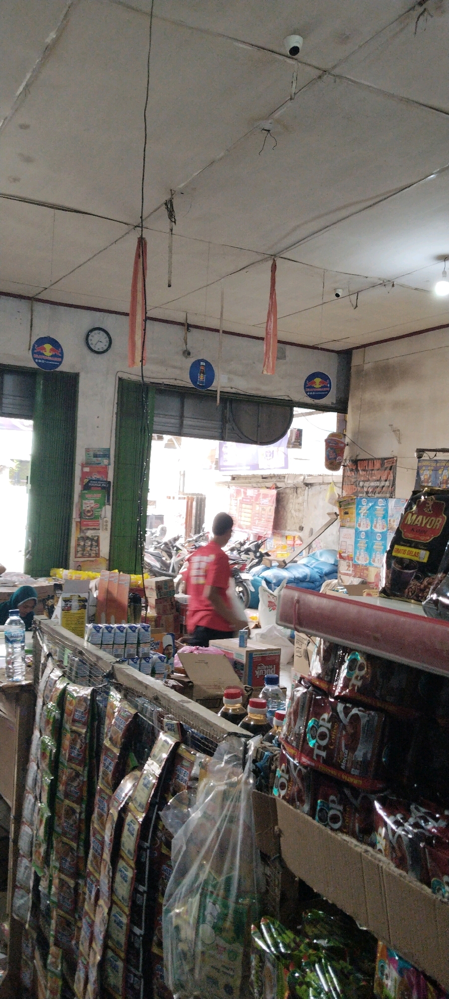 The image depicts the interior of a small convenience store, likely a "warung," in Bandar Jaya, Indonesia. The store is densely stocked with various goods, including packaged food items, drinks, and household supplies. The ceiling is made of suspended tiles and features exposed wiring, lighting fixtures, and two visible CCTV cameras. Red and white fabric streamers hang from the ceiling.
In the foreground, shelves are filled with neatly arranged packets of snacks and beverages, some still in their original packaging. Bottles of water and what appear to be soy sauce bottles are also visible on a counter. A person wearing a red t-shirt with a white design on the back is seen from behind, carrying a white plastic bag and walking away from the camera. Another person, partially visible on the left, is wearing a dark headscarf and appears to be interacting with the counter.
In the background, through an open doorway or entrance, a street scene is visible with several motorcycles parked. The store is dimly lit, with natural light entering from the front. Signs and advertisements are visible on the walls, some in Indonesian. The overall impression is one of a busy, local retail environment.