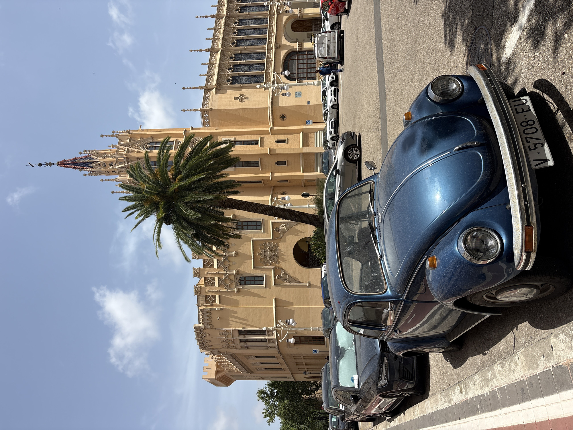 The image shows a street scene in Valencia, Spain, featuring a distinctive yellow building with Gothic-inspired architecture and a tall, ornate tower topped with a spire. In the foreground, a blue vintage Volkswagen Beetle is parked on the left side of the street. Several other cars are parked behind it, including a dark-colored Audi. A palm tree stands prominently in front of the building, casting a shadow on the street. The sky is blue with a few scattered clouds. The building appears to be a historic landmark or public institution.