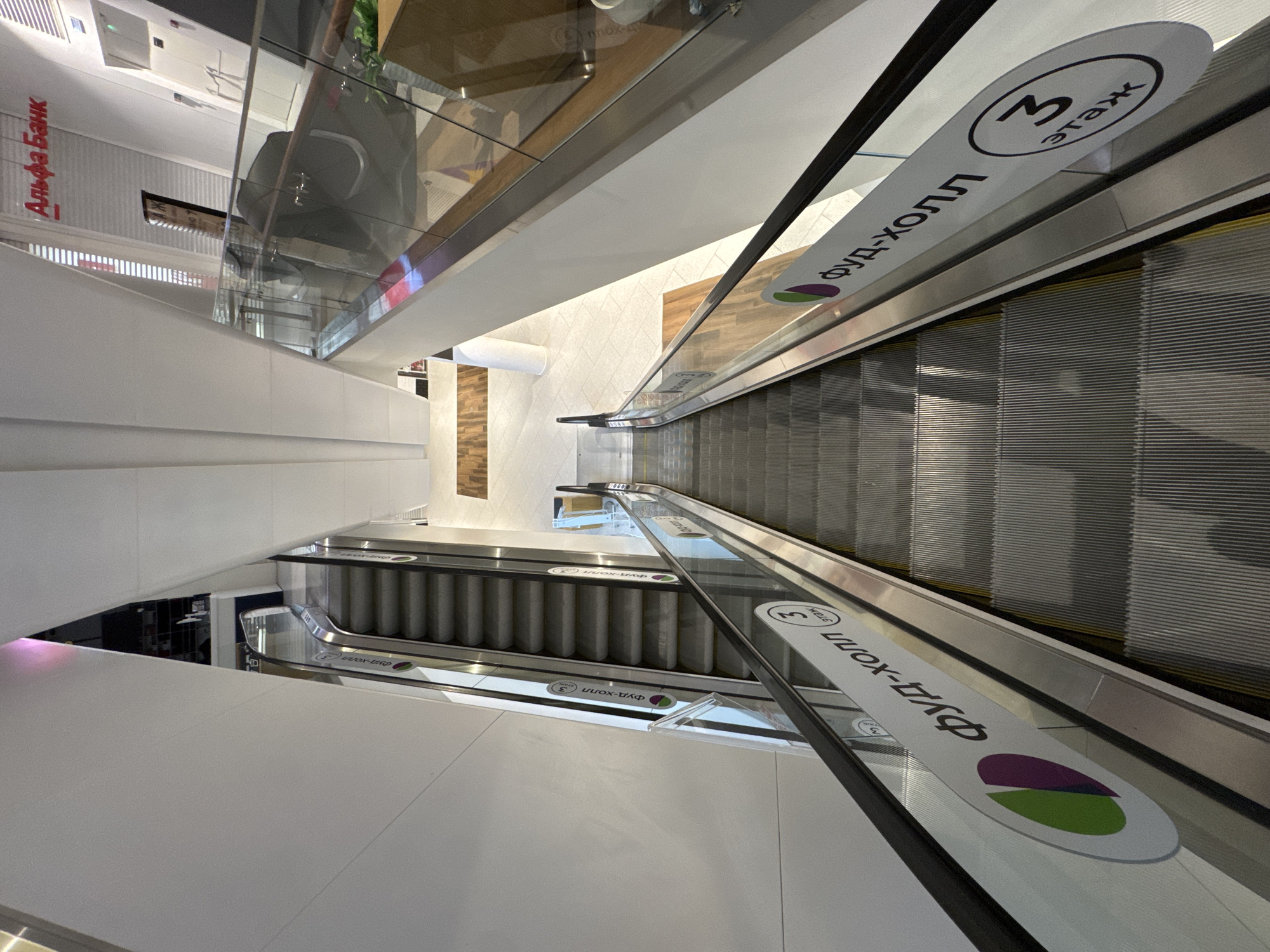 The image shows an escalator inside a modern building, likely a shopping mall or commercial center. The escalators have a signage indicating "Food Hall" on the 3rd floor, with the phrase written in Cyrillic, alongside a visual circular logo in purple and green. One side of the image also features a sign for "Alfa Bank", a large Russian private bank. The environment appears clean and well-lit, with white walls and neutral floor tiles. The escalators are oriented upwards, and the perspective is a high-angle shot looking down.