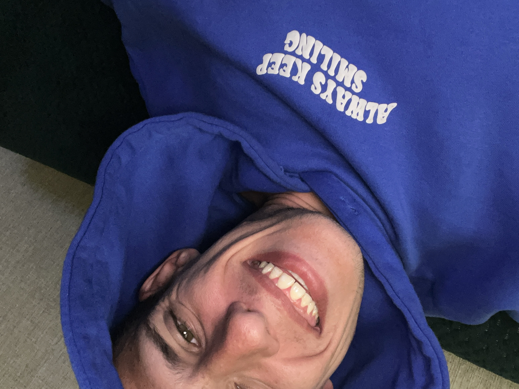 A selfie-style, close-up image displays a man wearing a blue hooded sweatshirt. The sweatshirt has the words "ALWAYS KEEP SMILING" printed on it in white. The man is smiling broadly, showcasing his teeth. His face is angled in the frame. The background includes a dark area and a lighter, textured surface. The location is not identifiable as Sofia, Bulgaria, based on visual cues in the image.