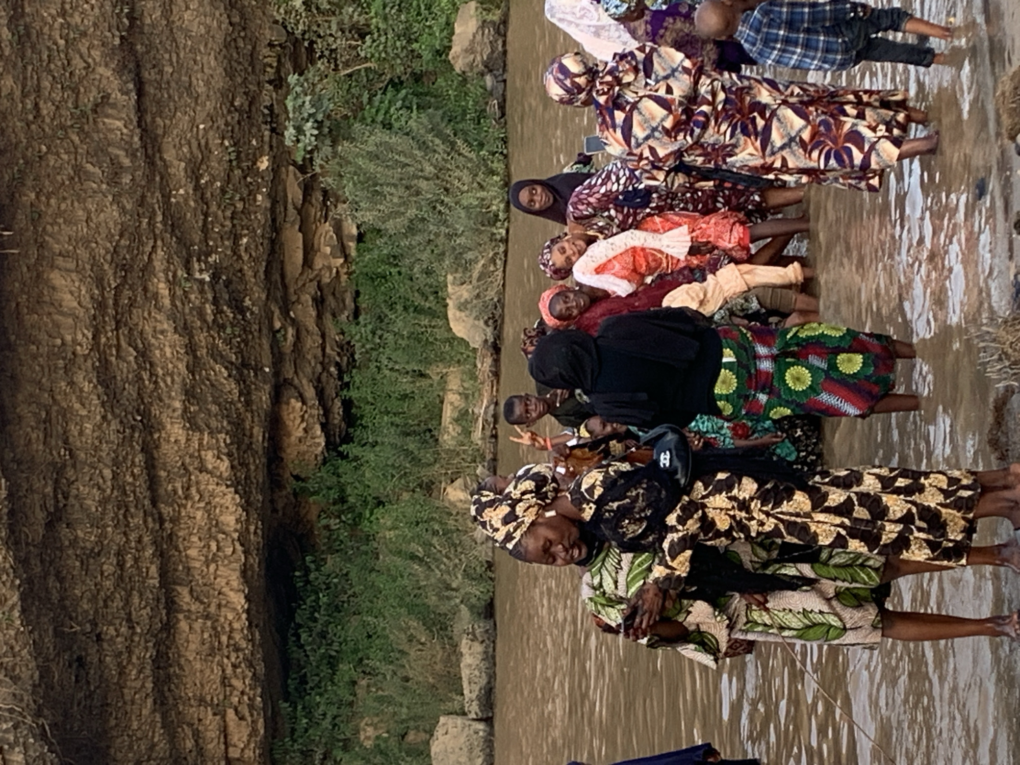 A group of people, mostly women and children, are standing in a body of water. The women wear a variety of colorful patterned fabrics. The location appears to be Kukum, Nigeria, indicated by the rock formation on the left. Some of the individuals are interacting with each other.