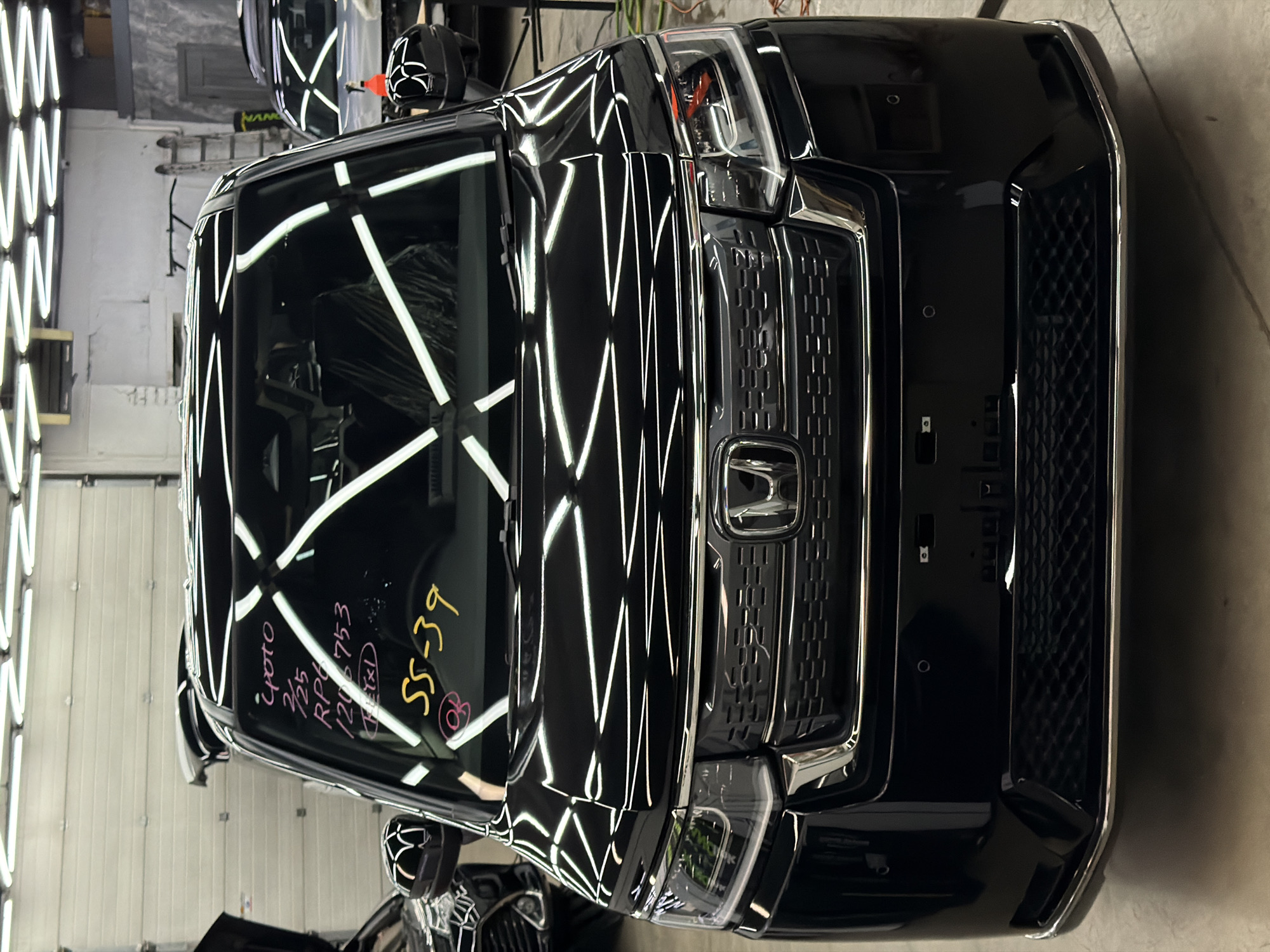 The media file displays a sleek, black car, likely a modern SUV or minivan, situated indoors within what appears to be a professional auto detailing or inspection facility. The image is oriented vertically, showing the car from a slightly elevated front-right perspective.
The primary subject is the car, gleaming with a highly reflective surface that showcases numerous overhead LED strip lights. These lights are arranged in a distinctive pattern, appearing as bright white 'X' shapes across the windshield and hood, characteristic of lighting setups designed to reveal paint imperfections or highlight a polished finish. The front of the car features a prominent chrome-bordered grille with a textured pattern and the silver Honda logo centrally placed. Modern headlights are visible, with intricate internal structures. The bumper also has textured black grilles.
No people are visible in the image. The setting is an indoor space, suggested by the structured lighting, painted walls (some appearing white or light gray, others concrete), and visible utility elements like pipes or cables in the background. The floor appears to be concrete.
Notable details include the immaculate, highly polished surface of the vehicle, indicating it might have recently undergone detailing, ceramic coating, or paint correction. The reflections of the overhead lights are sharp and undistorted, emphasizing the quality of the car's finish.
There is handwritten text visible on the windshield of the car, written in pink, yellow, and possibly red markers. The text reads:
* "Yoto" (pink)
* "2/25" (pink)
* "RP6 753" (pink)
* "120L" (pink, with a circle around 'L')
* "1x1" (pink, within a box)
* "SS-39" (yellow)
* A circled 'B' (red/pink)
These markings suggest internal tracking information, perhaps related to a work order, vehicle identification, or inspection details within the facility. The overall impression is one of a vehicle being prepared or inspected with meticulous care in a specialized environment.