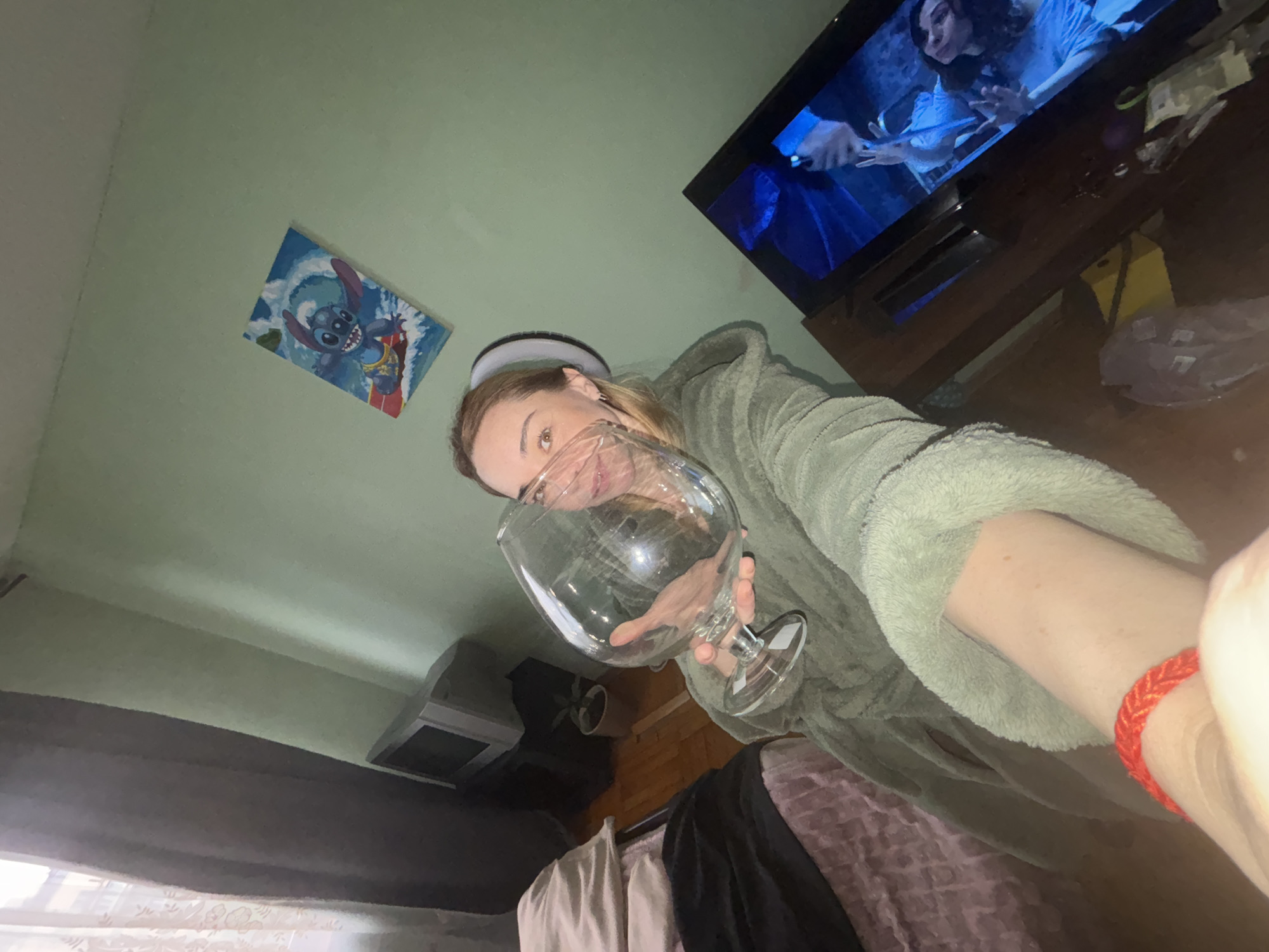 A woman in a green, hooded robe and a headband is holding a large, clear glass in front of her face. A television on a dark brown stand displays a scene from a film, while a "Stitch" drawing hangs on the light green wall. An older television set sits on the floor near the base of the wall. The floor is a medium brown wood. Clothing drapes over the bed. A red bracelet is on the wrist of the arm holding the camera.