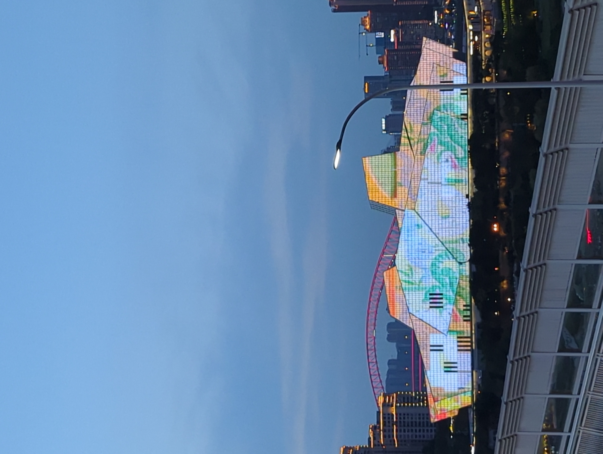 The image depicts a modern, architecturally unique building illuminated with vibrant, abstract patterns of light, situated in the Yuzhong District of China. The building's facade is a complex arrangement of geometric planes, covered in a grid of lights displaying a colorful projection that resembles foliage and abstract shapes. In the background, a prominent red suspension bridge arches across the cityscape, and further in the distance, numerous high-rise buildings are visible, some also illuminated. A streetlamp with a contemporary design stands in the foreground on the right side of the image, its light casting a subtle glow. The sky is a deep blue, indicative of dusk or twilight, with some faint clouds present. The overall scene conveys a sense of urban development and modern aestheticism, with the illuminated building serving as a focal point.