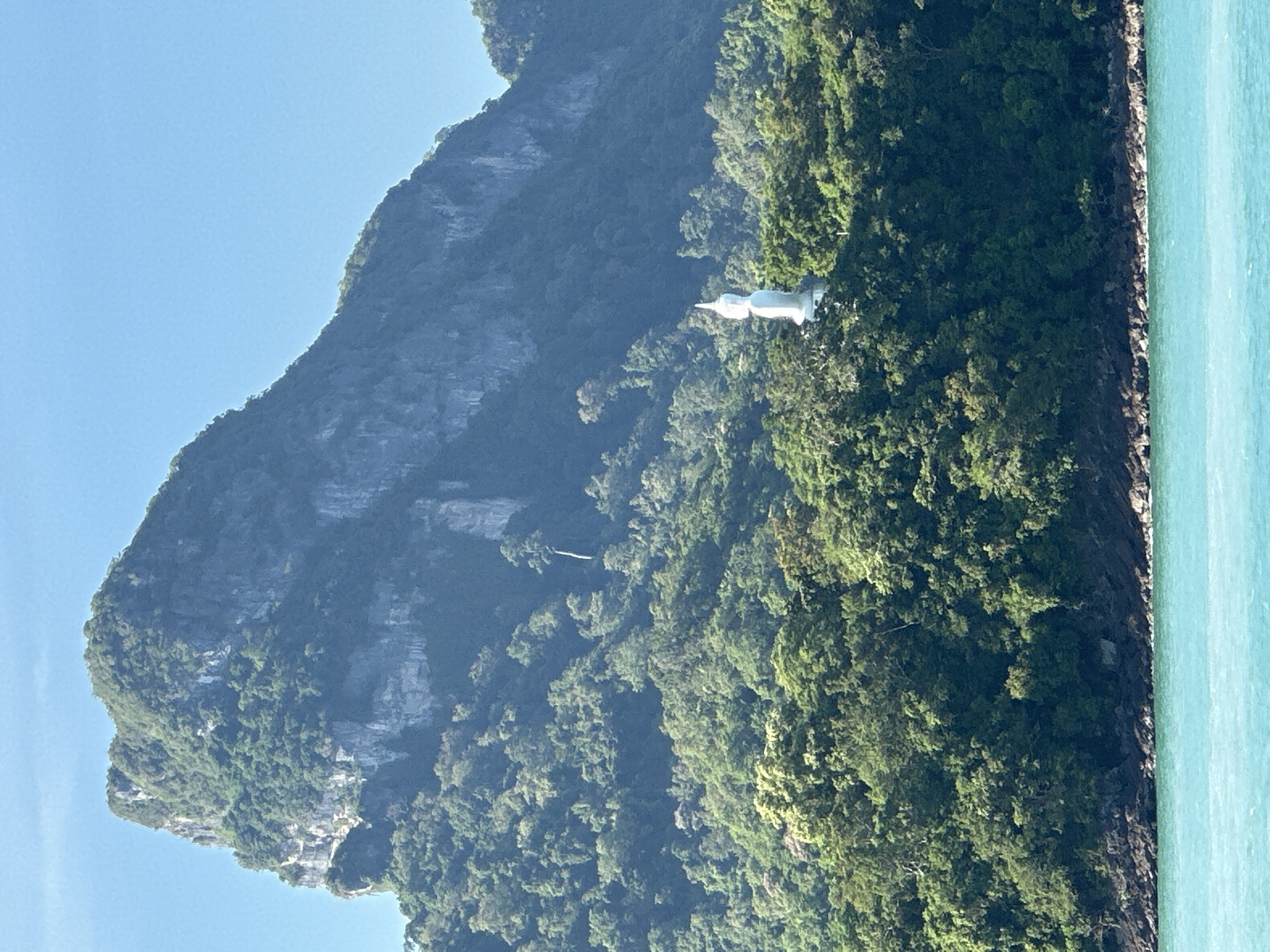The image shows a lush, green hillside dotted with trees, leading up to a large, rocky mountain peak under a clear blue sky. A white statue of a seated Buddha is visible on the hillside, partially obscured by the trees. In the foreground, a body of turquoise water laps at the rocky shoreline. This scene is likely located on an island near Don Sak, Thailand, a region known for its natural beauty and Buddhist sites.