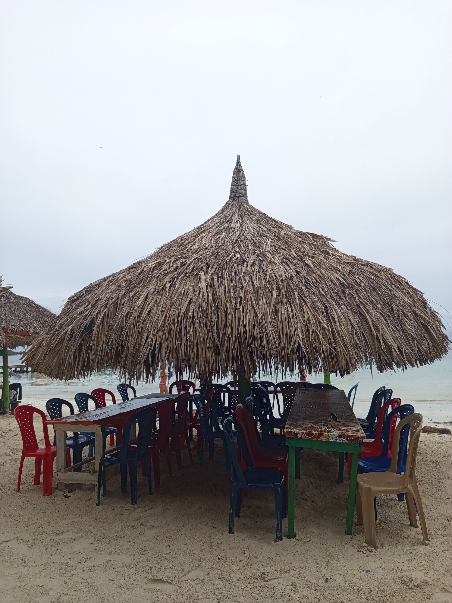 A thatched umbrella shades a long table and several plastic chairs on a sandy beach. The table is dark brown with a decorative edge, and it is supported by green legs. The chairs are a mix of red, blue, black, and tan. In the background, the ocean is visible, and beyond that, a wooden pier with some boats. A person wearing a pink bikini stands near the water. The sky is overcast. This scene appears to be a beachside dining area in a tropical location, possibly in a coastal city in Colombia given the mention of "Unknown, Colombia".