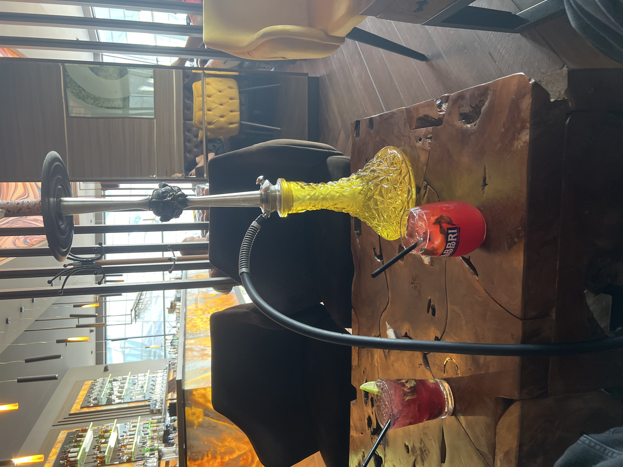 This media file captures an indoor scene, likely a bar or lounge, with a focus on a hookah and two cocktails. The image is taken from a slightly elevated perspective, looking down and across a rustic wooden table.
The main subjects are a prominent yellow, textured glass hookah with a silver stem and a black hose, positioned on a unique live-edge wooden table. Next to the hookah are two red or pink cocktails in clear glasses, each with a black straw. One cocktail has a lime wedge as a garnish. The word "CAMPARI" is clearly visible on the side of one of the cocktail glasses.
The setting appears to be an upscale and modern establishment, possibly in Burgas, Bulgaria. In the background, there are dark upholstered chairs or booths, and reflections of yellow tufted chairs can be seen in mirrored or glass surfaces. The ceiling features large windows or a glass roof, allowing natural light to illuminate the space, suggesting it is daytime. Modern, linear lighting fixtures hang from the ceiling. Further in the background, a well-stocked bar area with numerous bottles arranged on shelves is visible.
The overall activity depicted is one of relaxation and socializing, with patrons enjoying drinks and shisha. The wooden table has a distinct, carved, and uneven surface, adding a natural and artisanal touch to the interior design. The ambiance conveyed is sophisticated and comfortable.