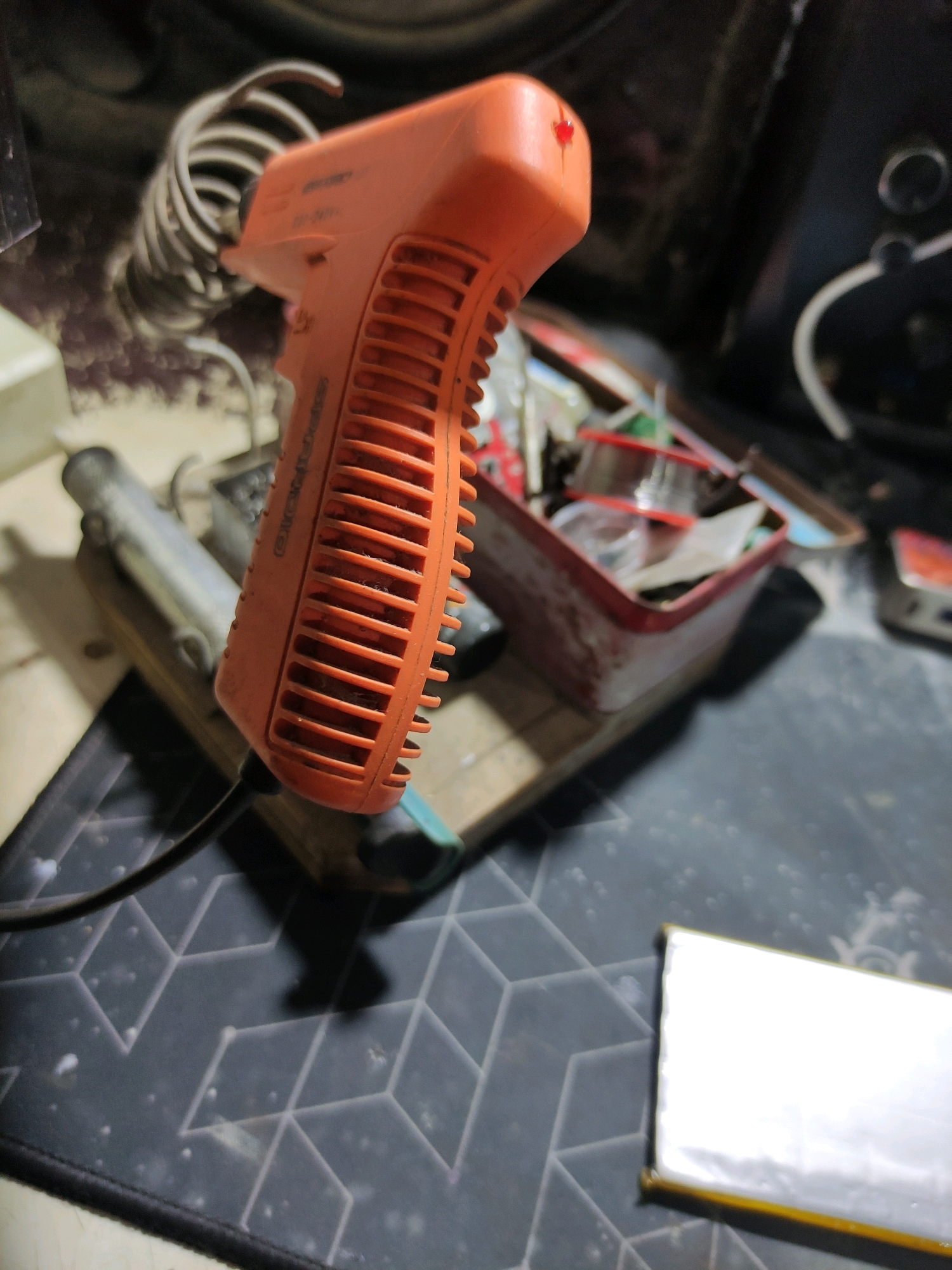 An orange soldering iron with a coiled cord rests on a wooden platform. In the background, a red-rimmed container holds spools of wire and other small items. The scene appears to be an indoor workspace, possibly a home or a small workshop, located in Bulukumba, Indonesia. The overall lighting is dim, suggesting it might be evening or a poorly lit area. There are no people visible in the image.