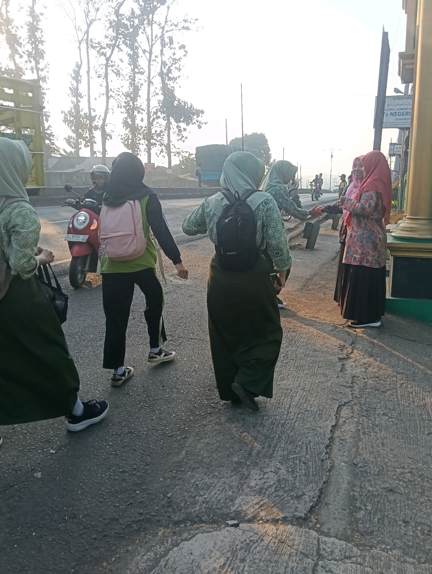 In Adinuso, Indonesia, on a sunny day, several young women in Islamic attire walk along a paved road. One woman in the foreground wears a light green hijab and a patterned top, while another next to her is in a dark hijab and a green shirt. A third woman, also in a light green hijab, faces away from the camera. Two women in the background are also visible. A red scooter is parked on the side of the road. A signpost for KEMENTERIAN AGAMA and ISLAMIC JUNIOR HIGH SCHOOL is visible on the right side of the image, indicating a school vicinity. The scene captures a moment of daily life in a semi-urban Indonesian environment, with a backdrop of trees and distant structures.
